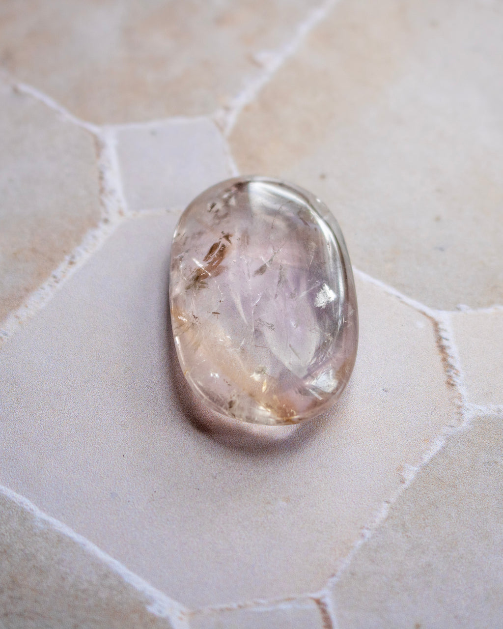 A polished oval cabochon gemstone with a milky pale lavender and beige color, containing tiny inclusions, resting on a light beige hexagonal tile surface.