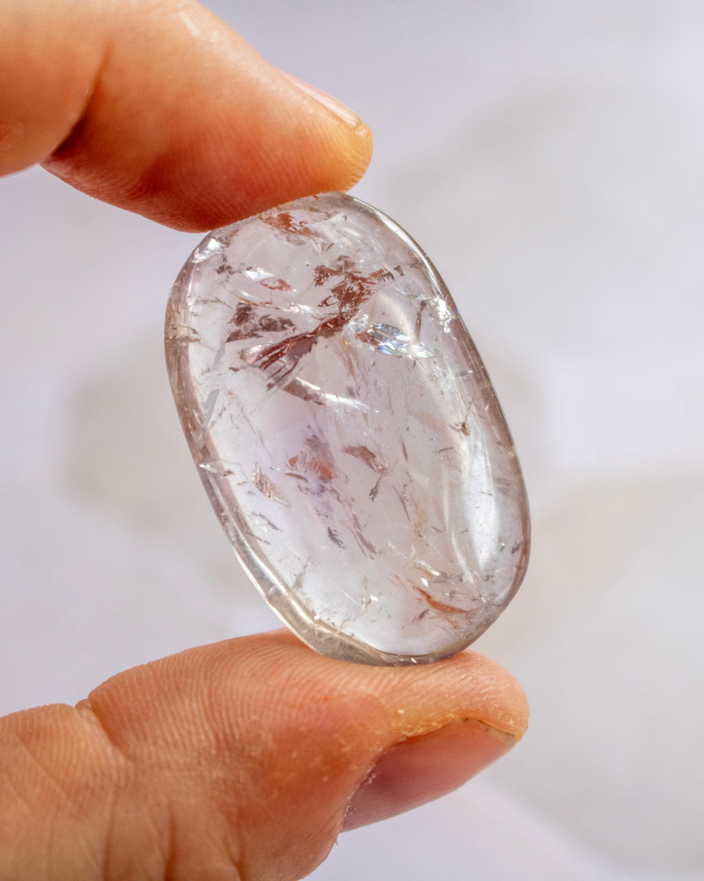A hand holding a polished, teardrop-shaped clear gemstone with visible brown and white inclusions inside, set against a soft, out-of-focus light background.
