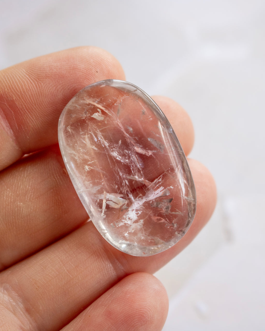 A hand holding a polished, oval-shaped clear crystal gemstone with visible internal inclusions and striations against a bright white background.