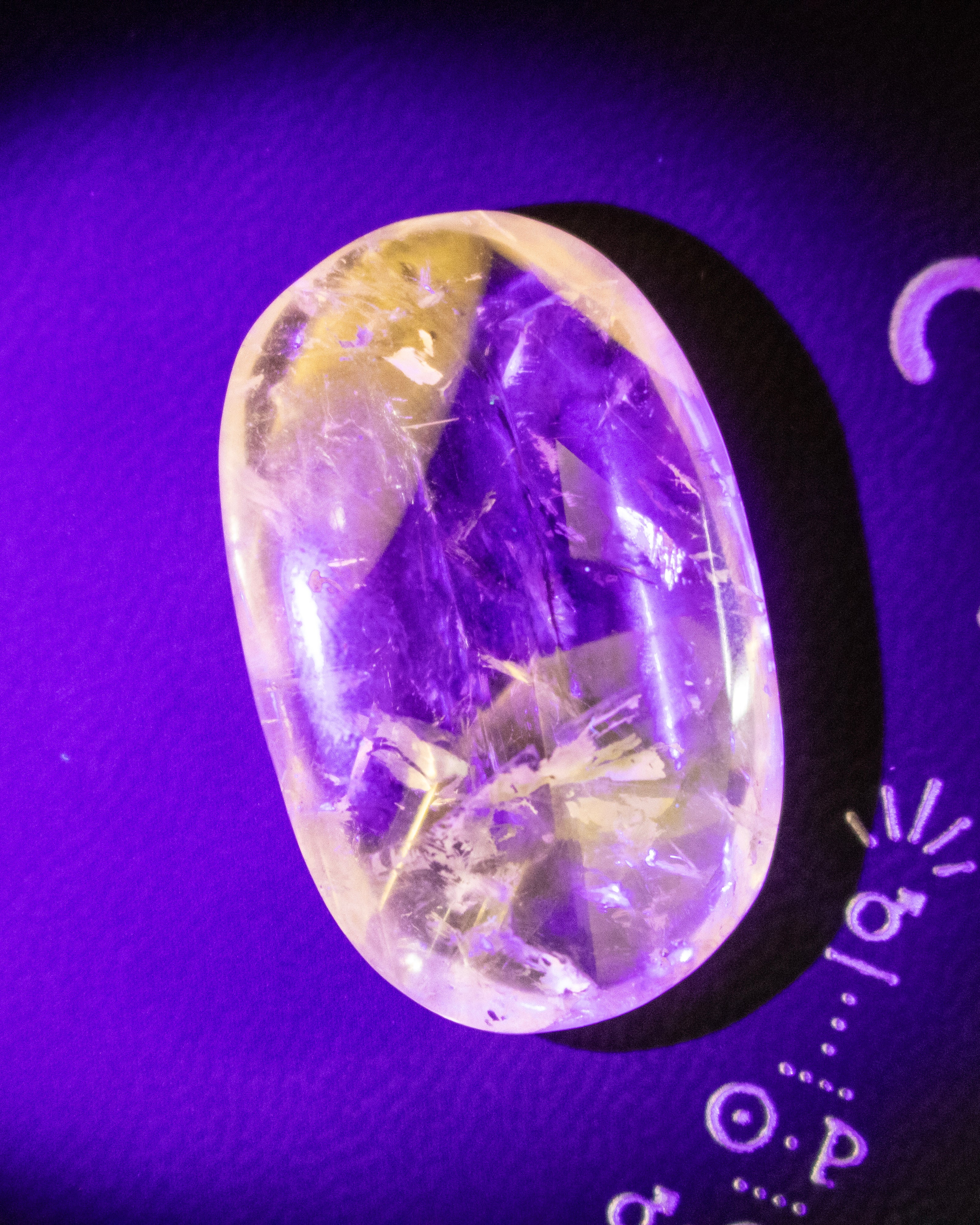 A polished oval gemstone cabochon with a purple and yellow internal pattern, displayed on a purple background with a small white scale marker.