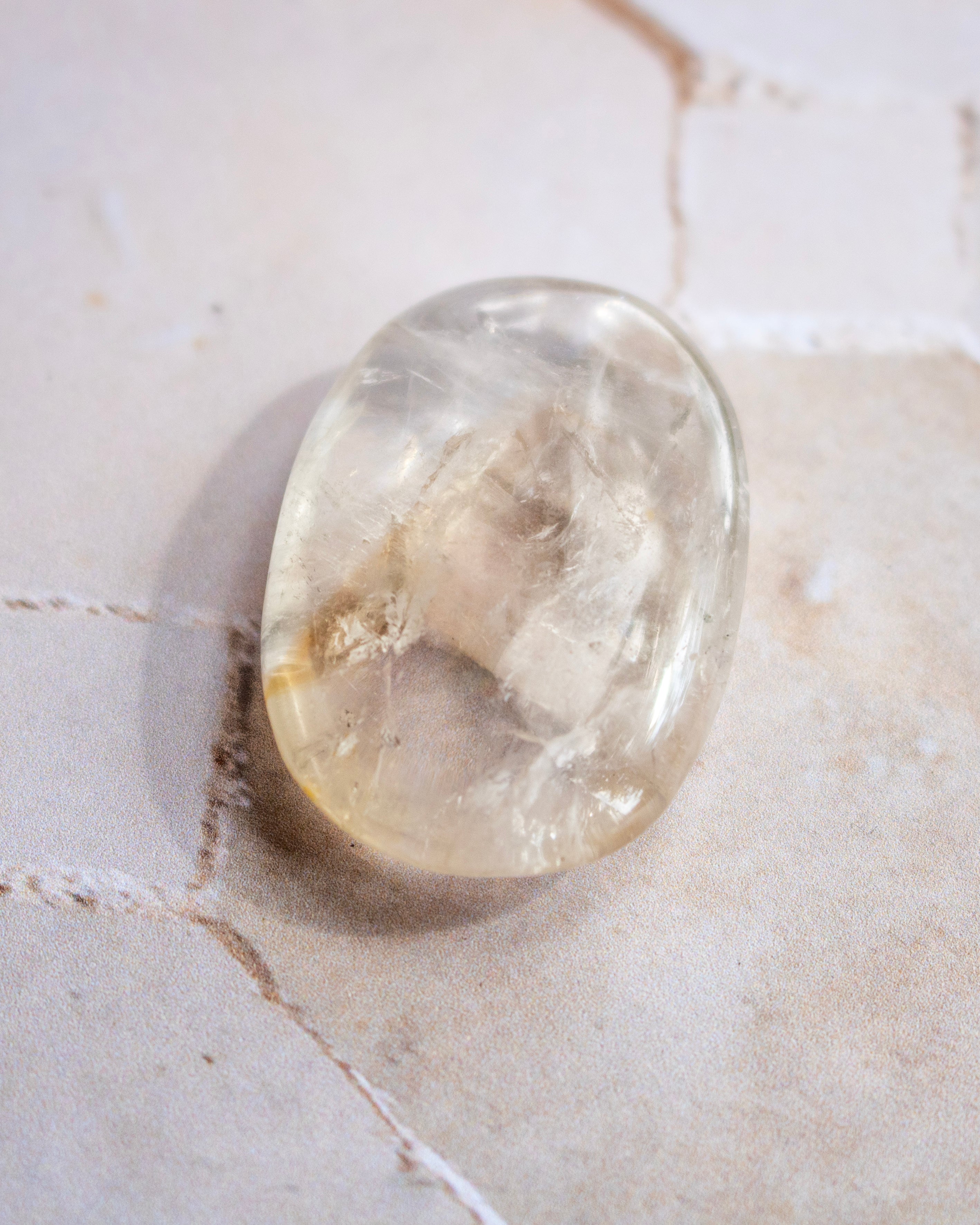 A polished, oval-shaped clear gemstone with natural internal inclusions, resting on a light beige stone surface.