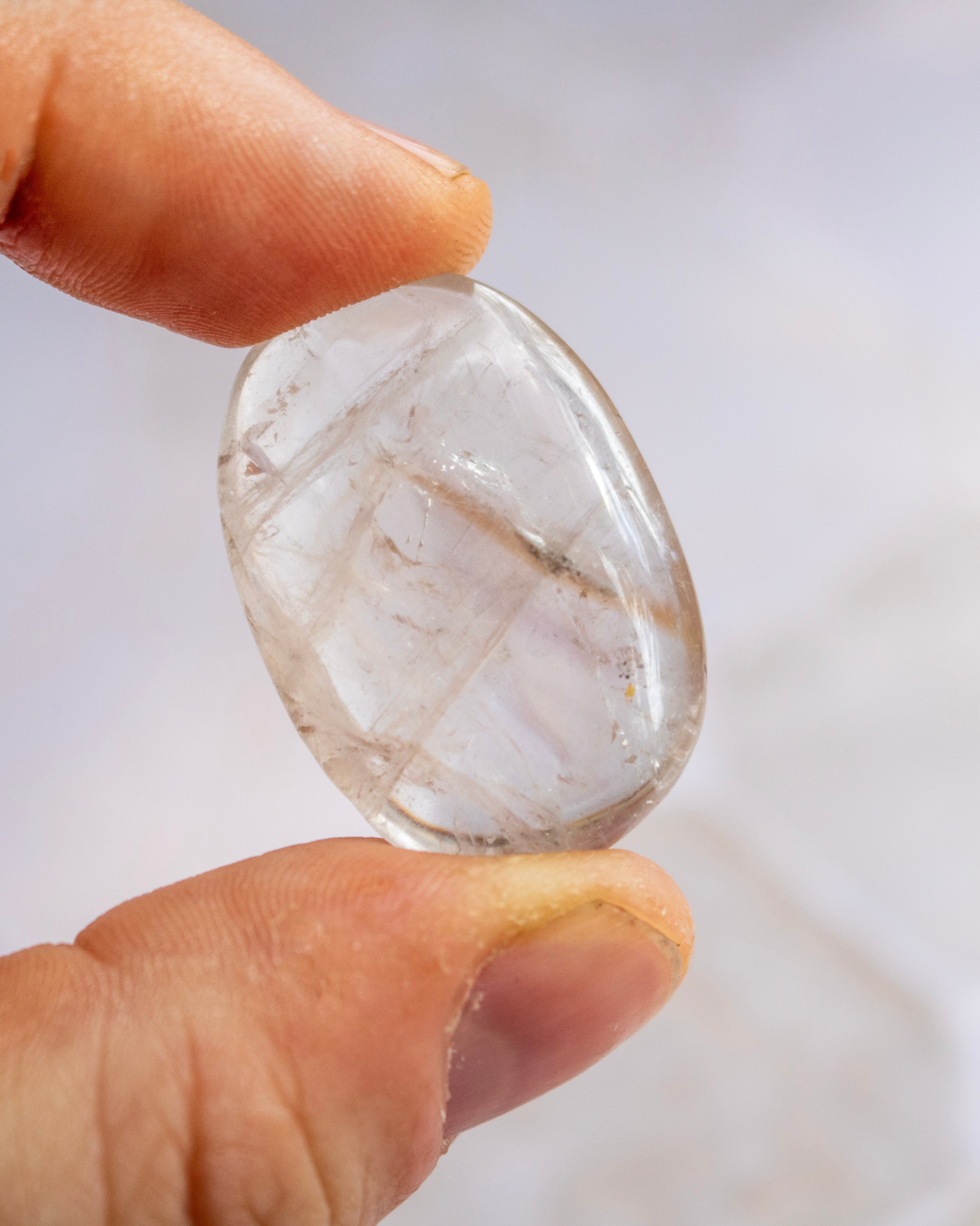 A hand holding a small, polished, teardrop-shaped clear gemstone with natural inclusions, shown against a soft, out-of-focus light background.