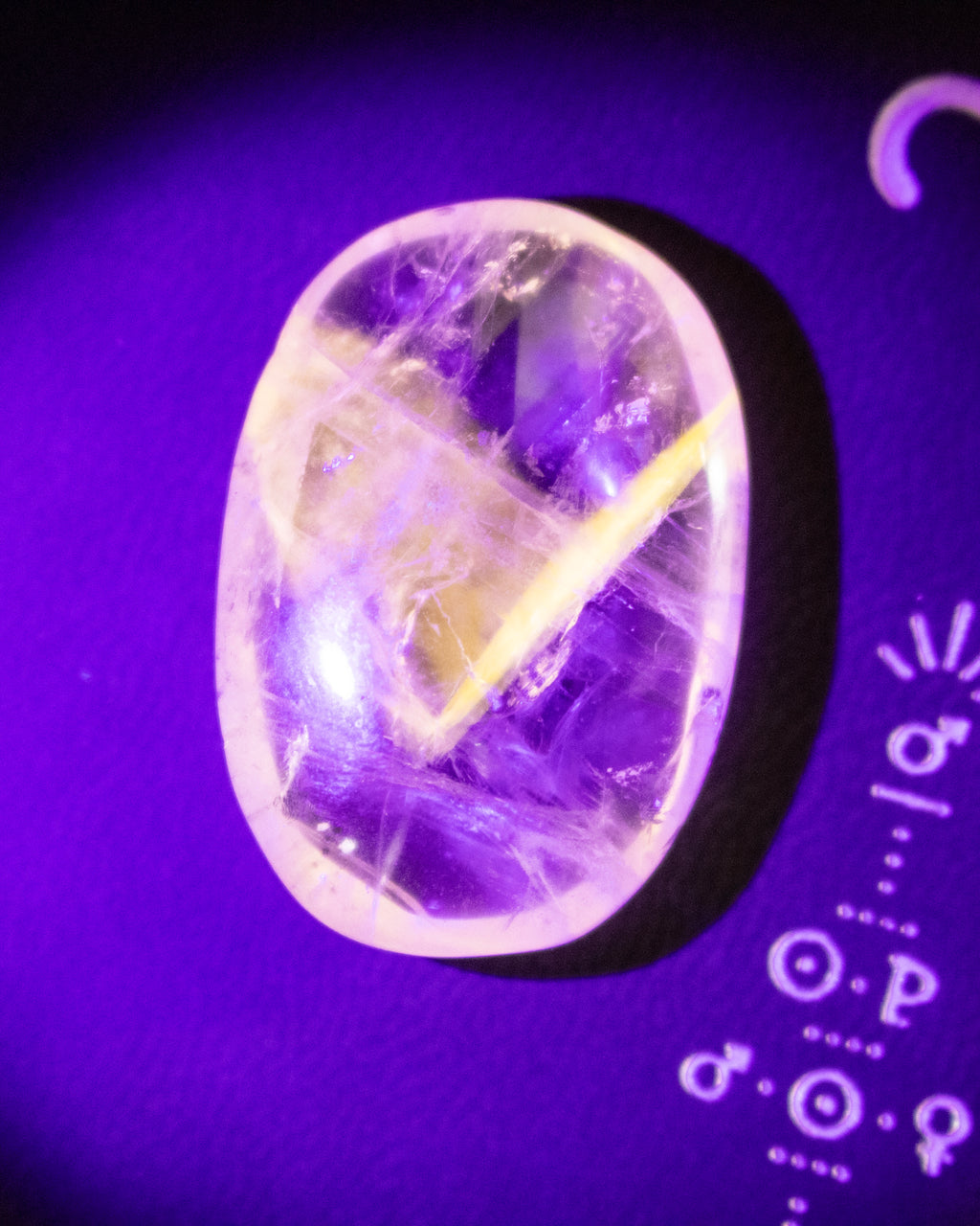 A polished oval gemstone cabochon with a clear and purple matrix, featuring a yellow starburst pattern, photographed under purple light on a dark background with astrological symbols.