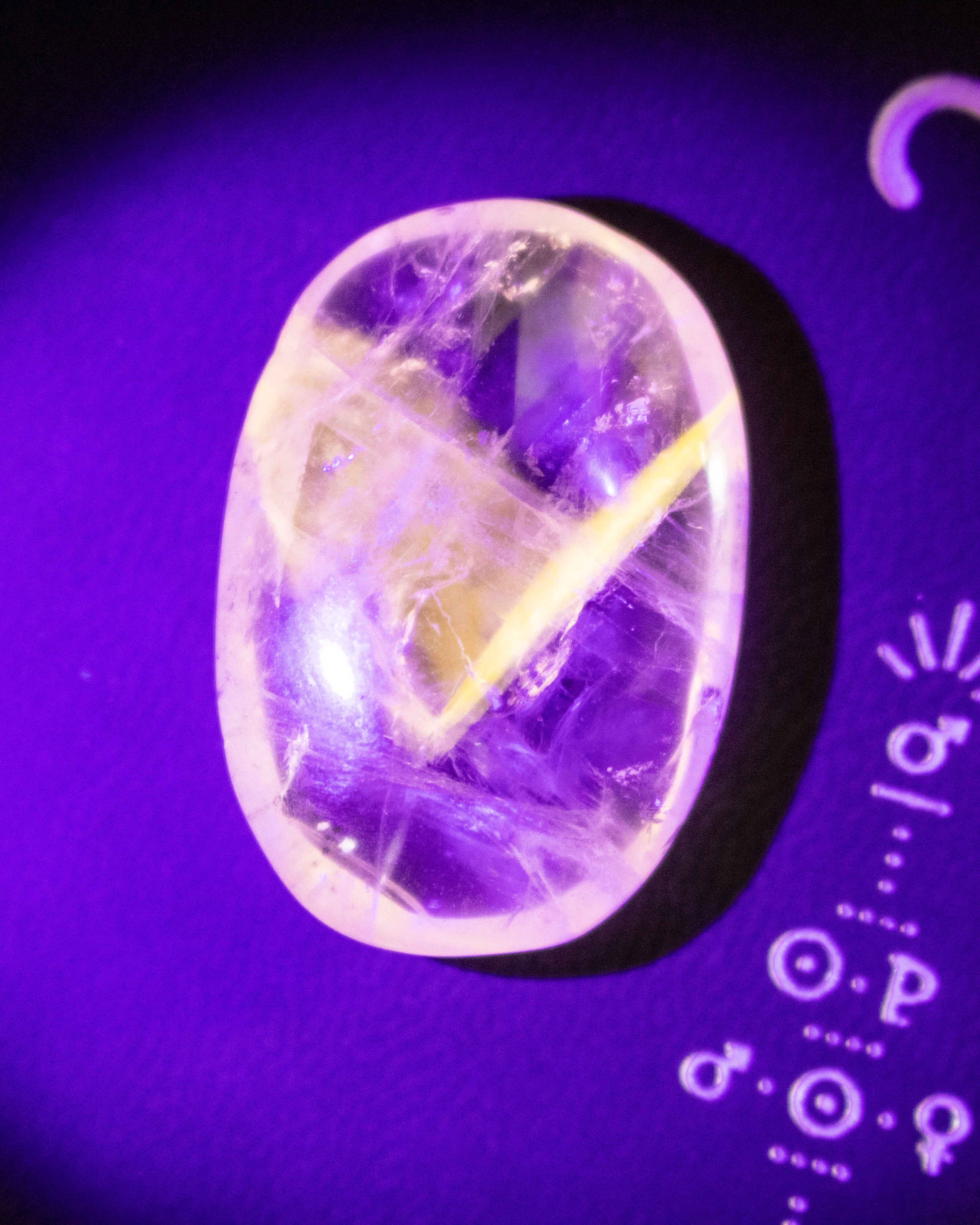A polished oval gemstone cabochon with a clear and purple matrix, featuring a yellow starburst pattern, photographed under purple light on a dark background with astrological symbols.