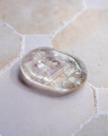 A polished, oval-shaped clear quartz crystal stone resting on a light beige stone surface.