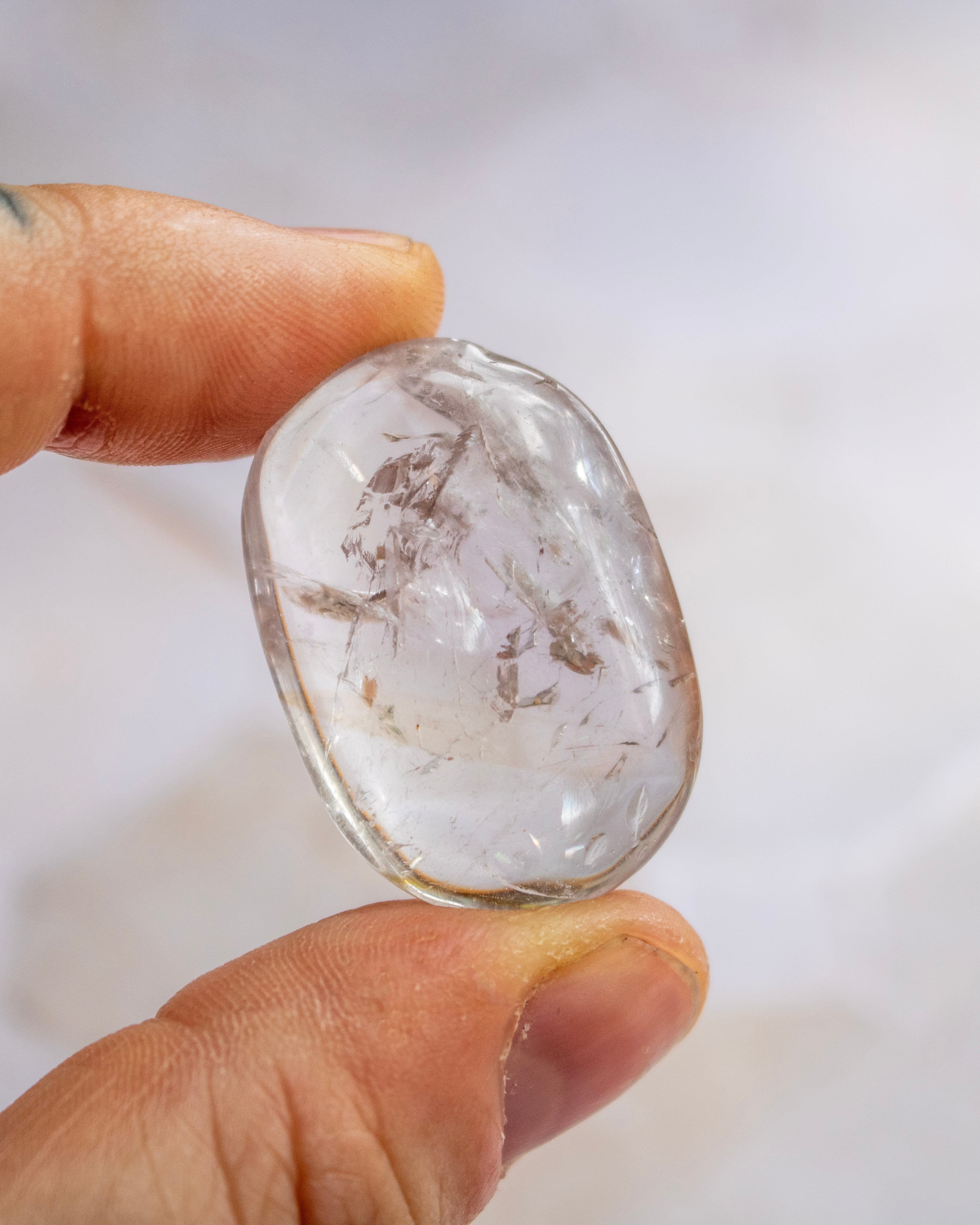 A hand holding a polished, oval-shaped clear gemstone with visible internal inclusions and a soft gray gradient background.