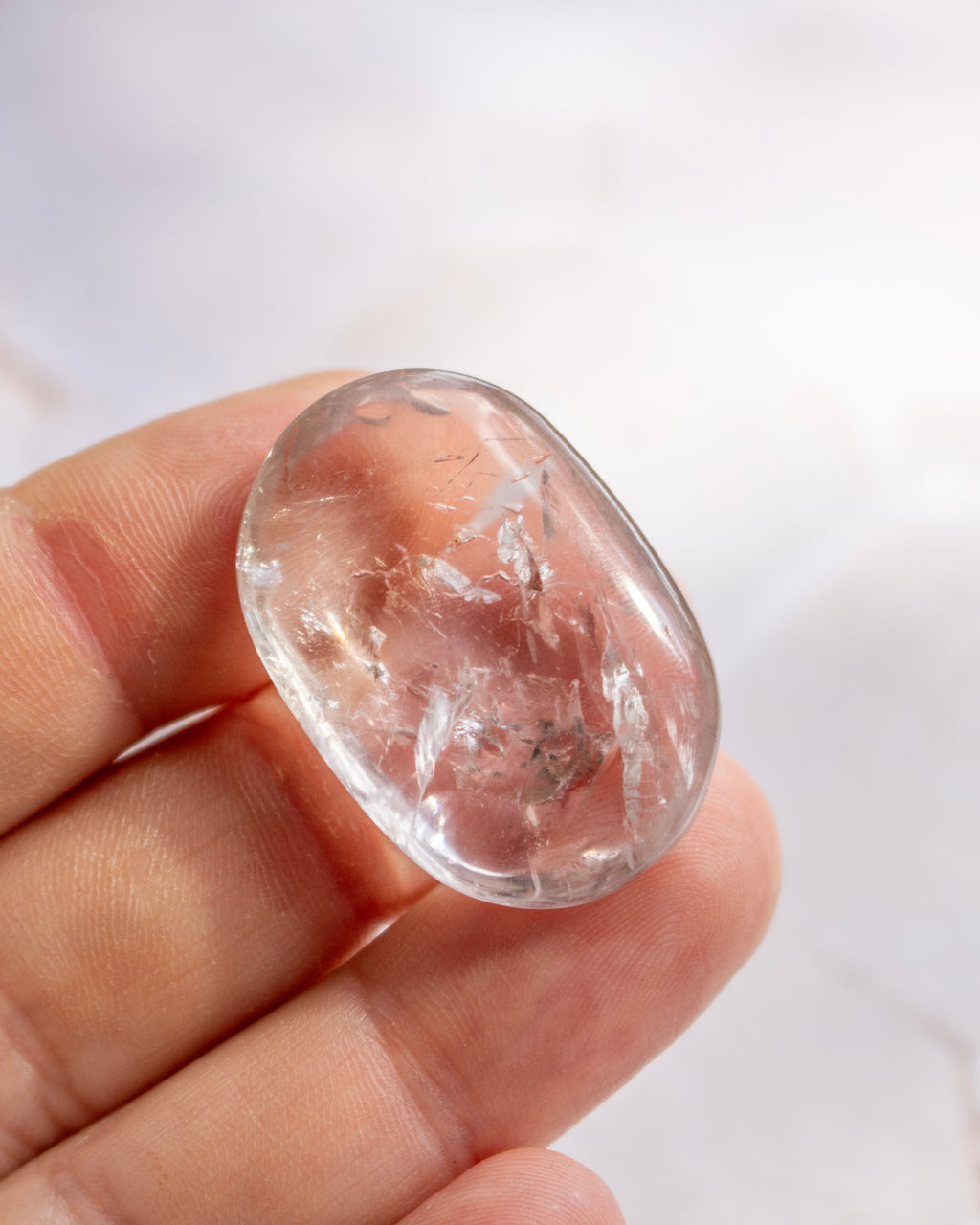 A hand holding a polished, oval-shaped clear quartz gemstone with visible internal inclusions against a soft, light background.