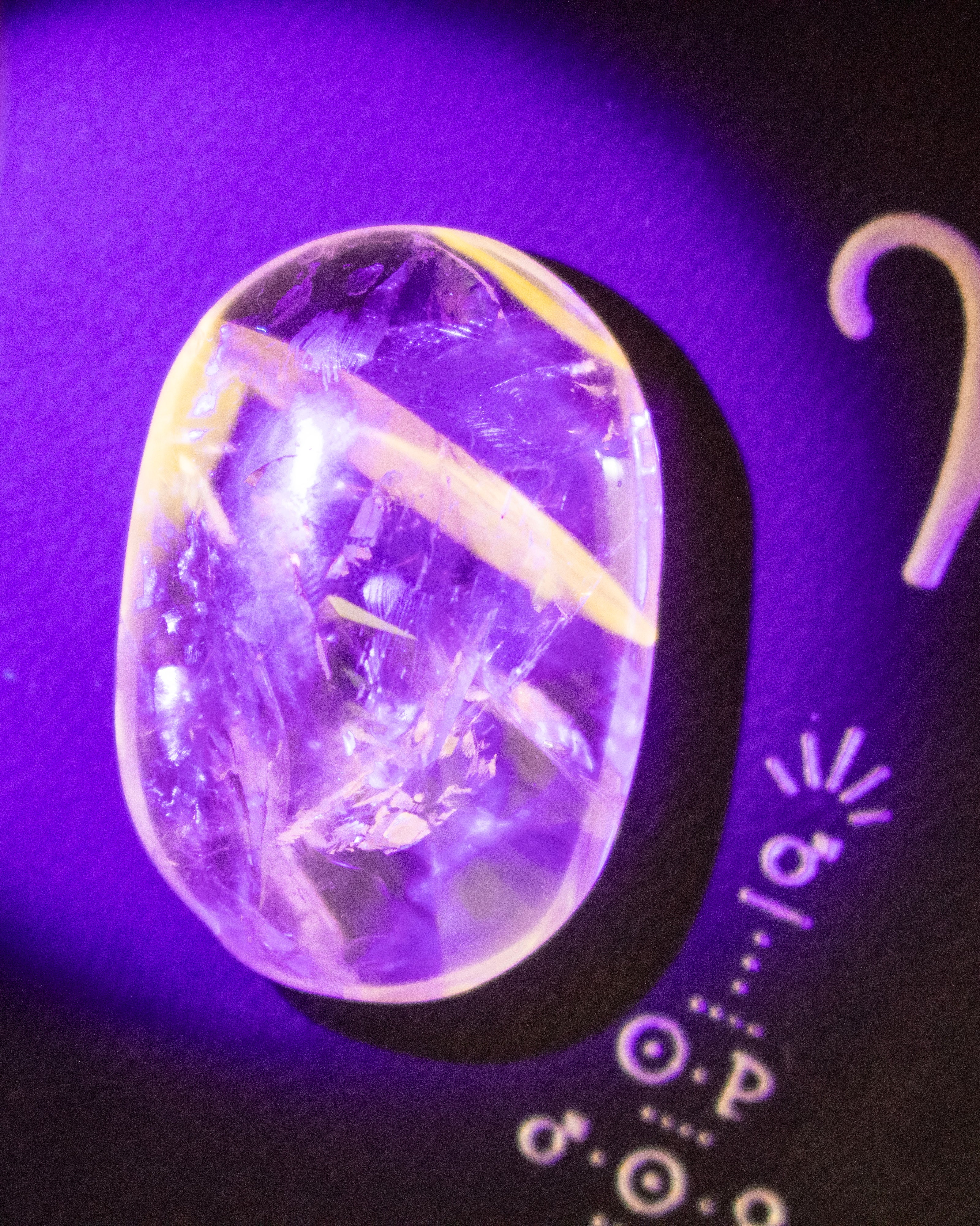 A polished oval clear gemstone with visible internal inclusions, photographed under purple light on a dark surface with white decorative markings.
