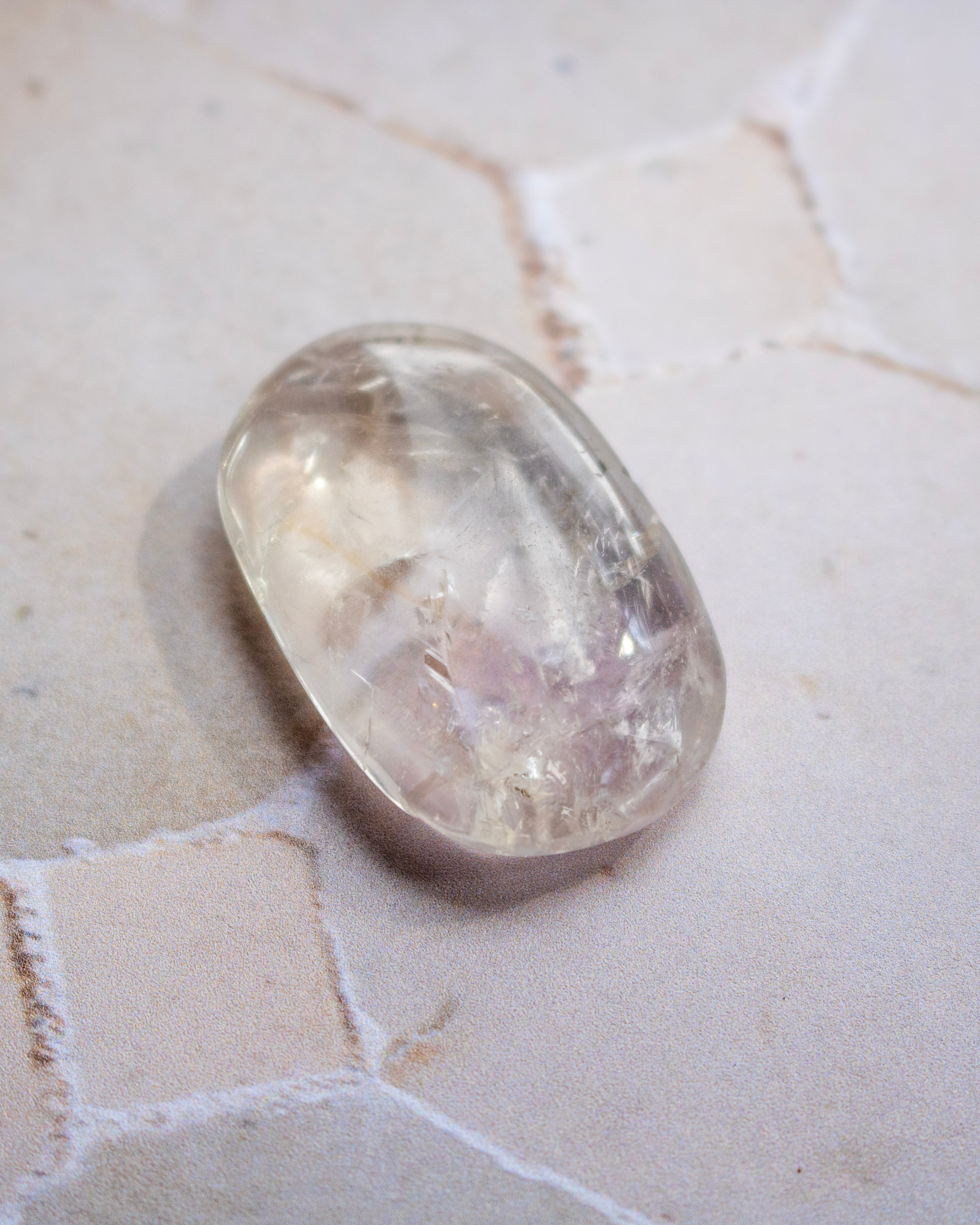 A polished oval-shaped clear gemstone with natural internal inclusions, resting on a light beige textured stone surface.