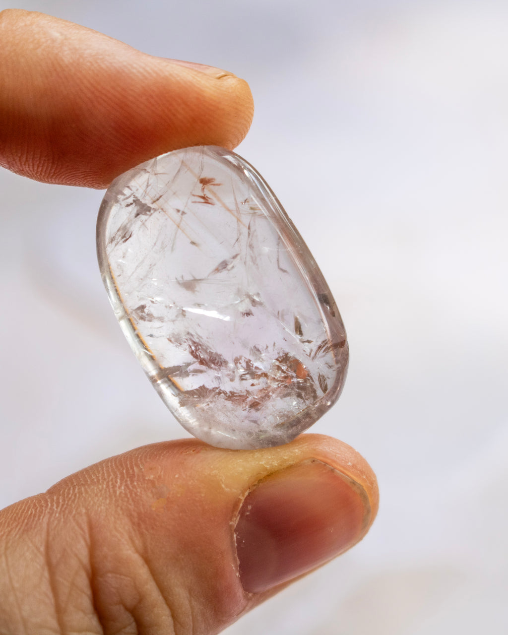 A close-up of a hand holding a polished, rounded clear gemstone with visible brown and white inclusions against a soft white background.