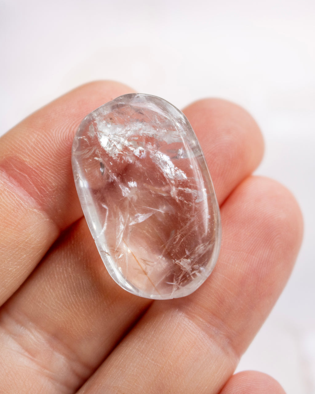 A small, polished oval clear gemstone with internal crystalline structures, held between a person's fingers against a soft white background.