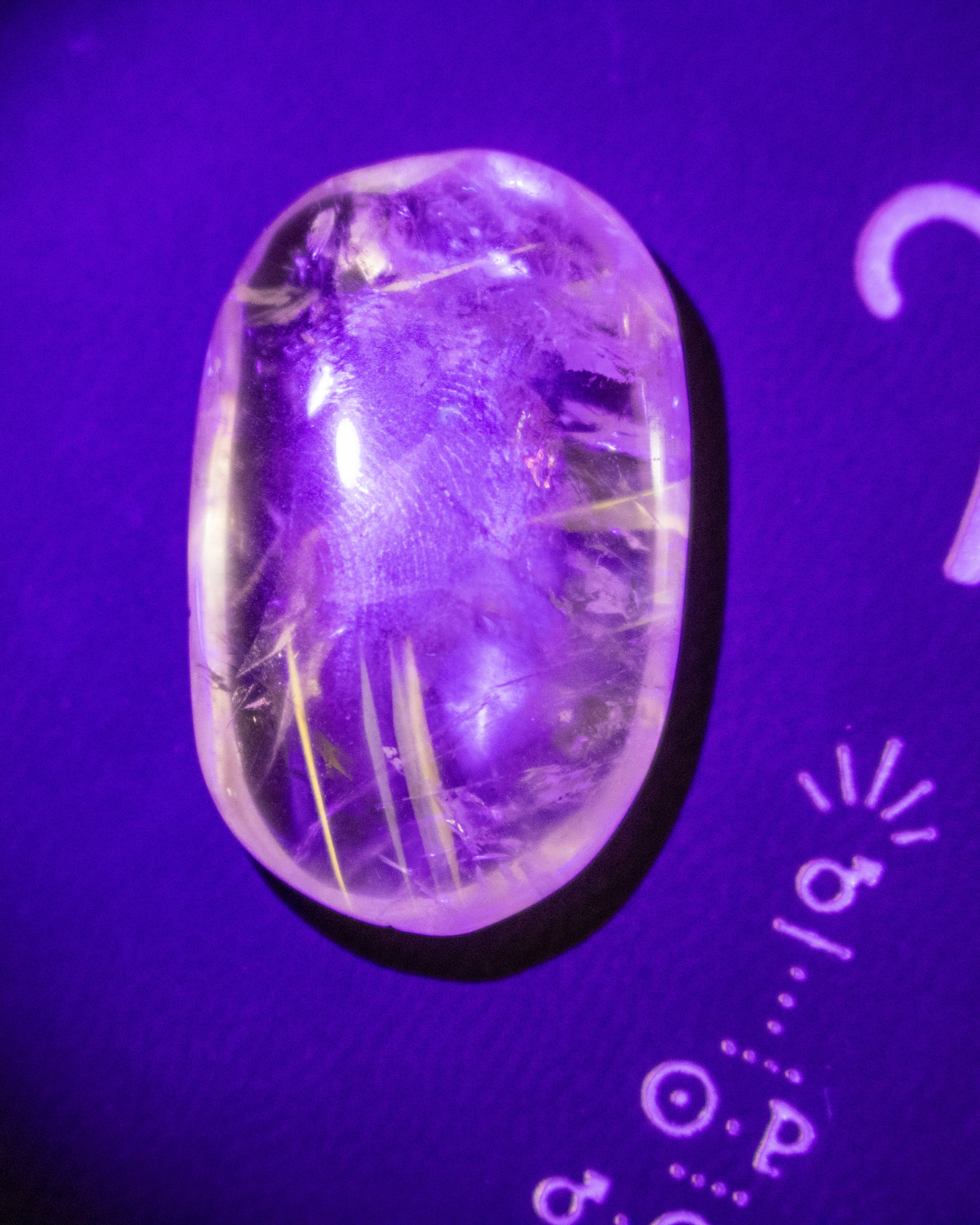 A polished oval clear gemstone with fine inclusions, shown under purple light on a purple background with printed measurement markings.