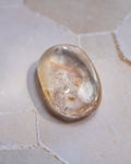 A polished, smooth oval-shaped clear gemstone with natural inclusions, resting on a light beige stone surface.