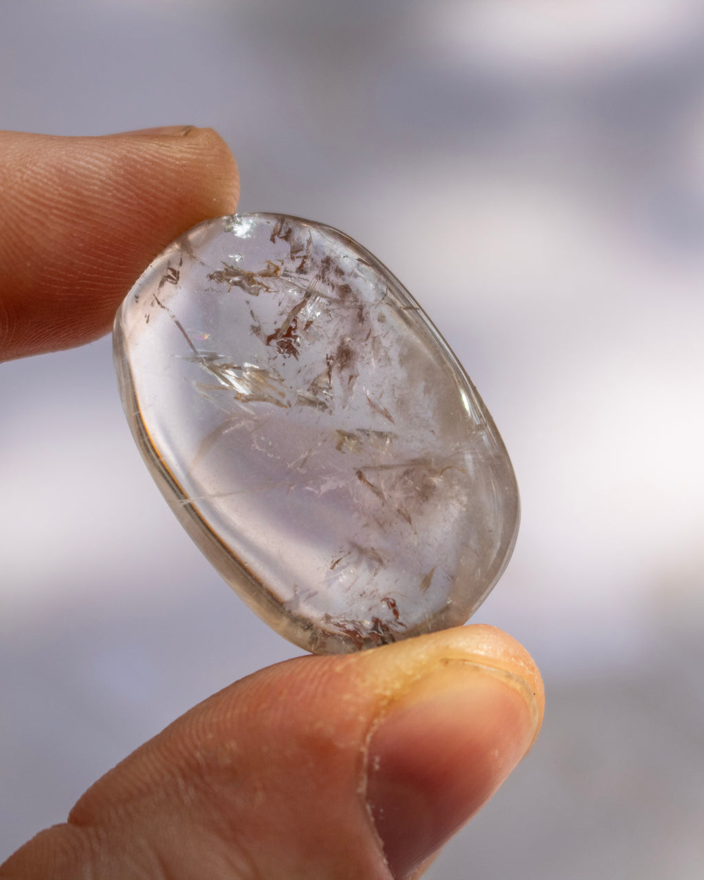 A hand holding a polished, teardrop-shaped clear gemstone with natural inclusions and brownish flecks inside, set against a soft, out-of-focus light gray background.