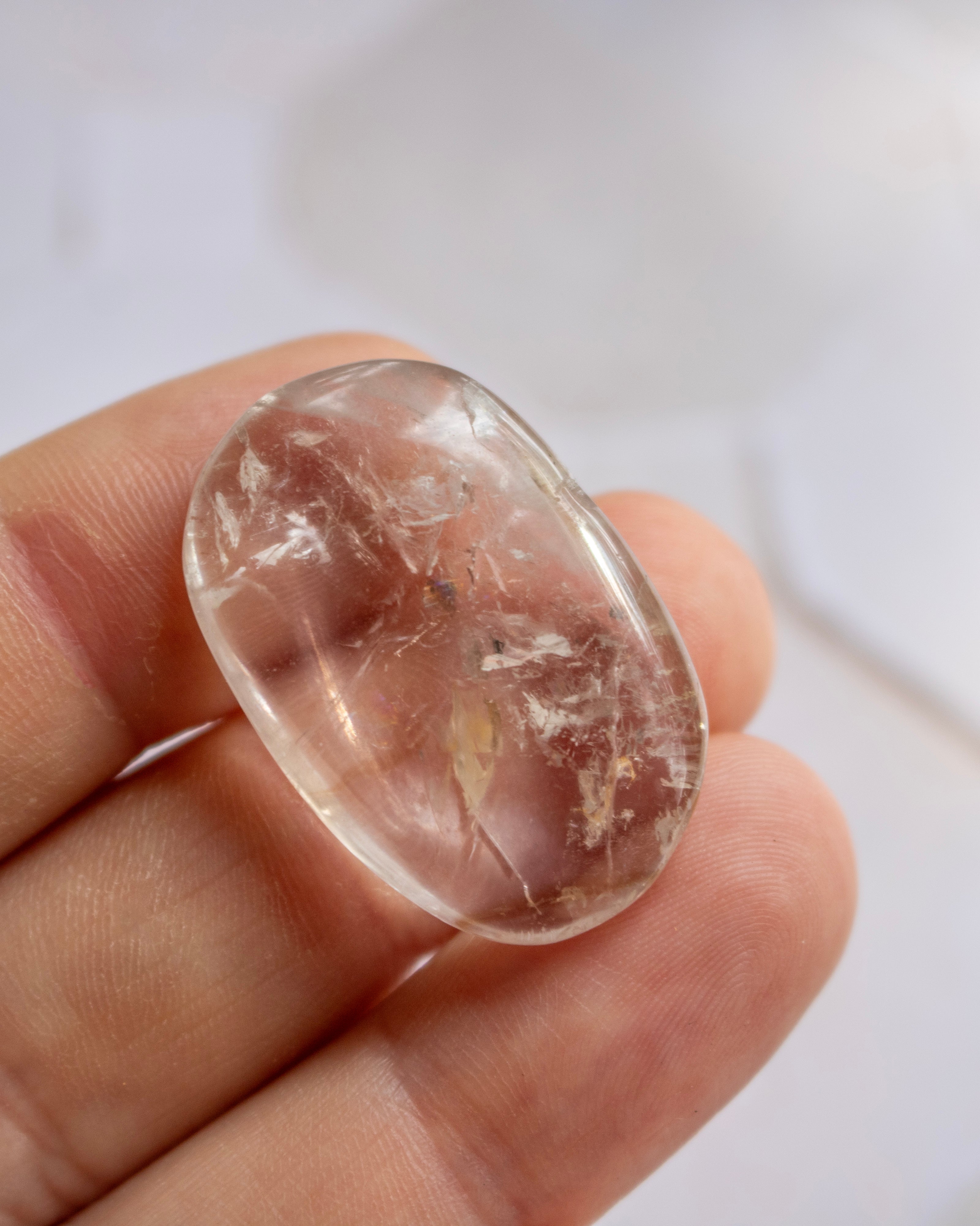 A hand holding a polished, rounded clear gemstone with internal inclusions, shown close-up against a soft white background.