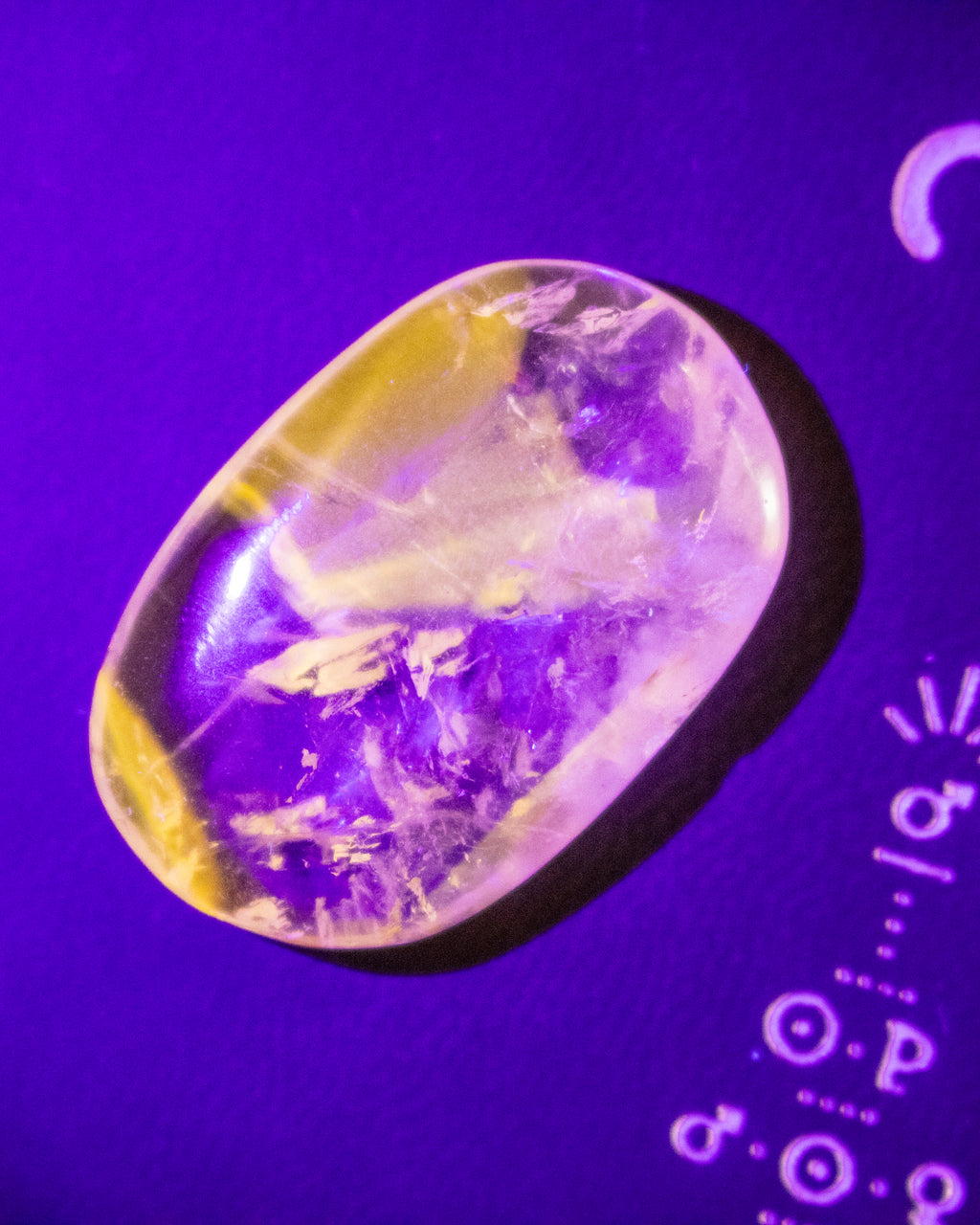 A polished oval gemstone with a yellow and purple banded pattern, displayed on a vibrant purple background with white lettering partially visible on the right.