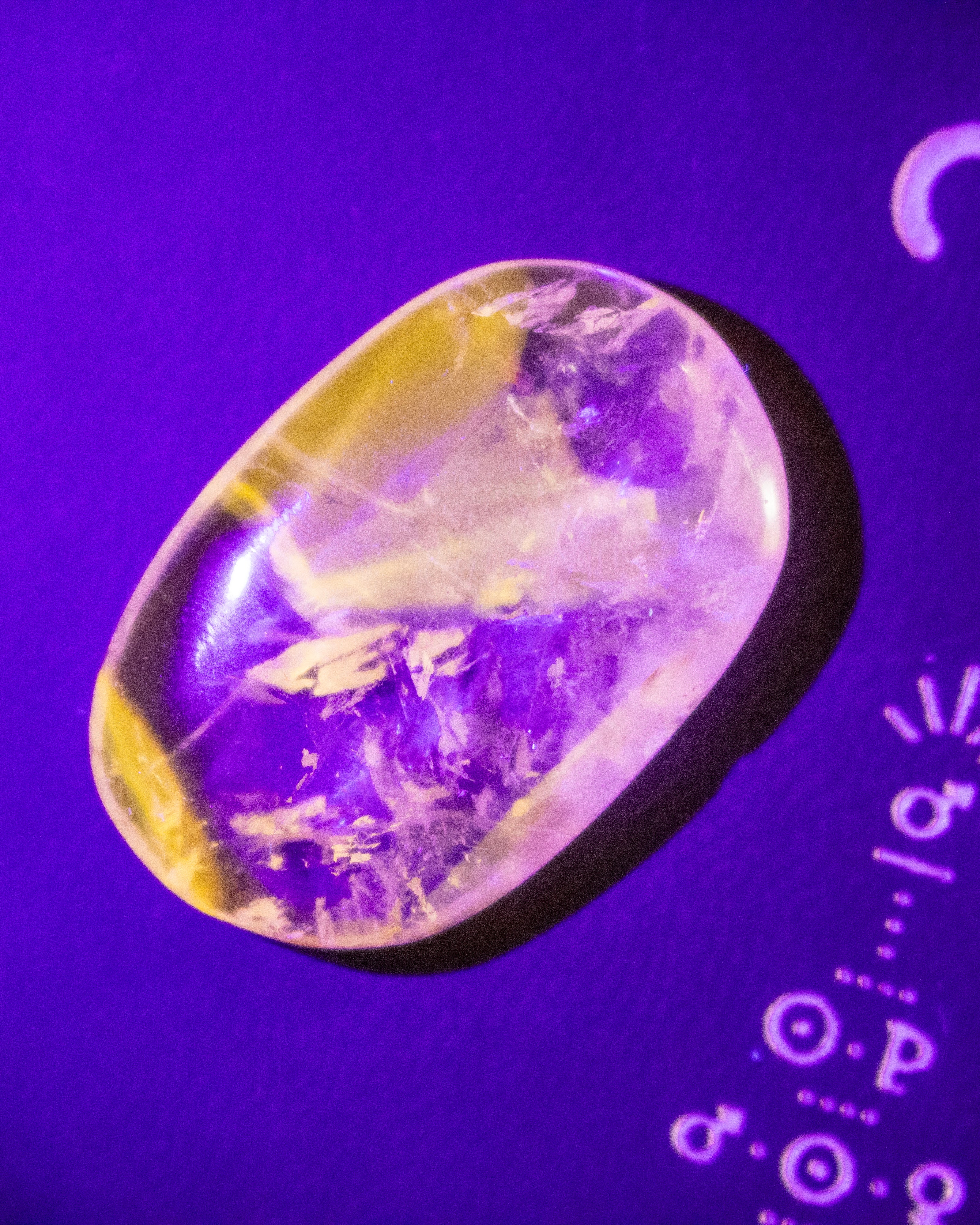 A polished oval gemstone with a yellow and purple banded pattern, displayed on a vibrant purple background with white lettering partially visible on the right.