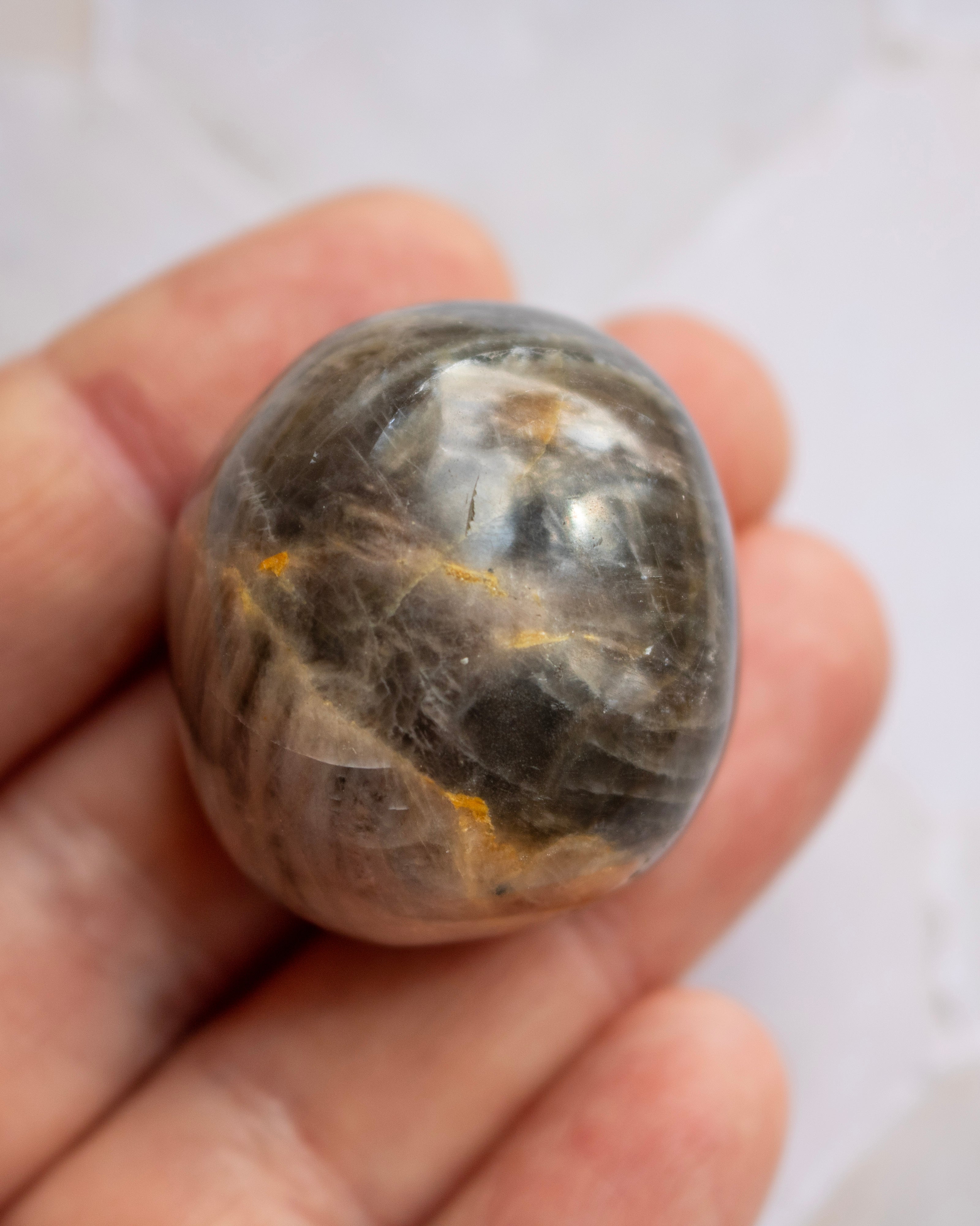 A polished oval gemstone with a smoky gray and brown body and visible golden-yellow inclusions, held in a person's hand against a light background.