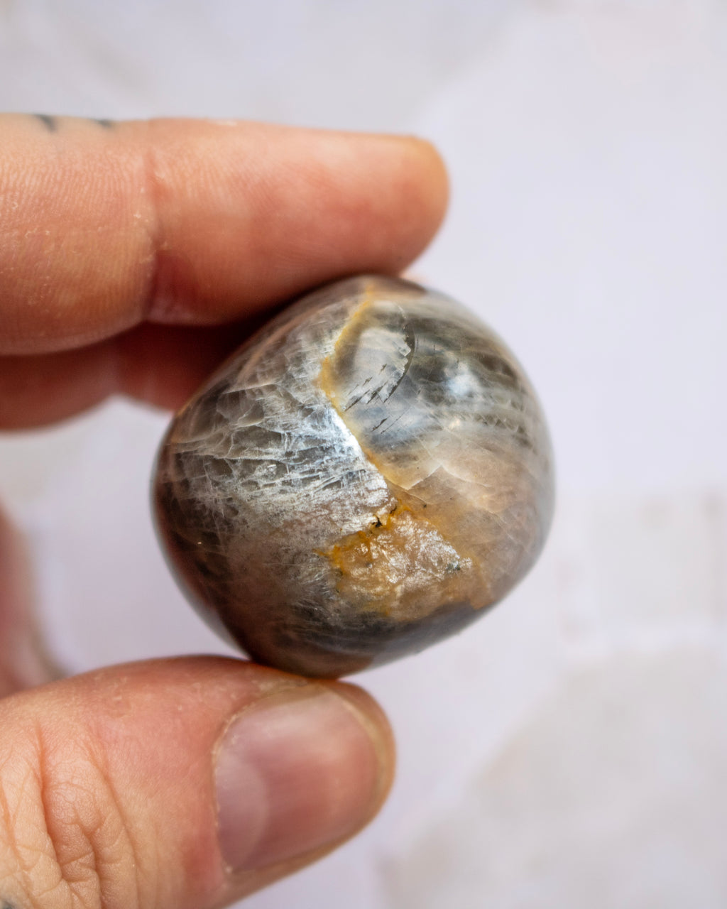 A close-up of a hand holding a polished stone or mineral specimen with a rounded, oval shape. The stone has a mottled appearance with gray, brown, black, and metallic gold veining.