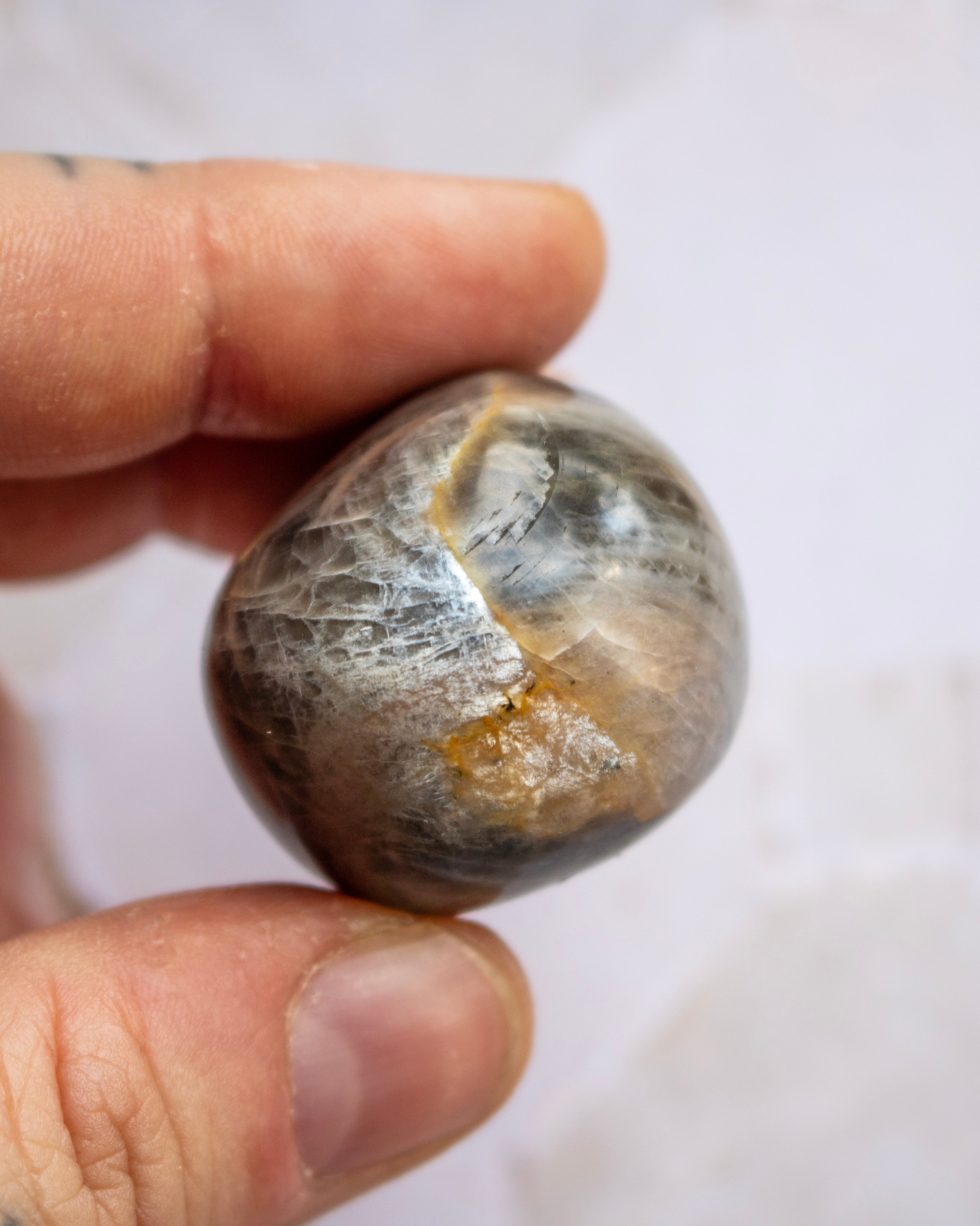 A close-up of a hand holding a polished stone or mineral specimen with a rounded, oval shape. The stone has a mottled appearance with gray, brown, black, and metallic gold veining.