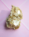 A rough, metallic gold crystal cluster with silver-toned facets, photographed on a soft pink background.