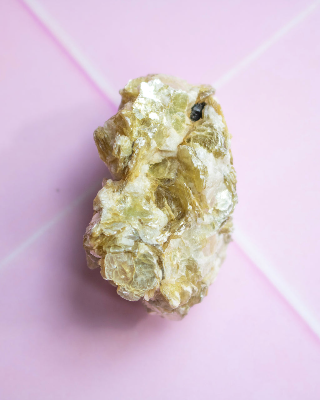 A raw, rough mineral specimen with a bubbly, crystalline texture in pale yellow-green and beige tones, photographed on a light pink background.