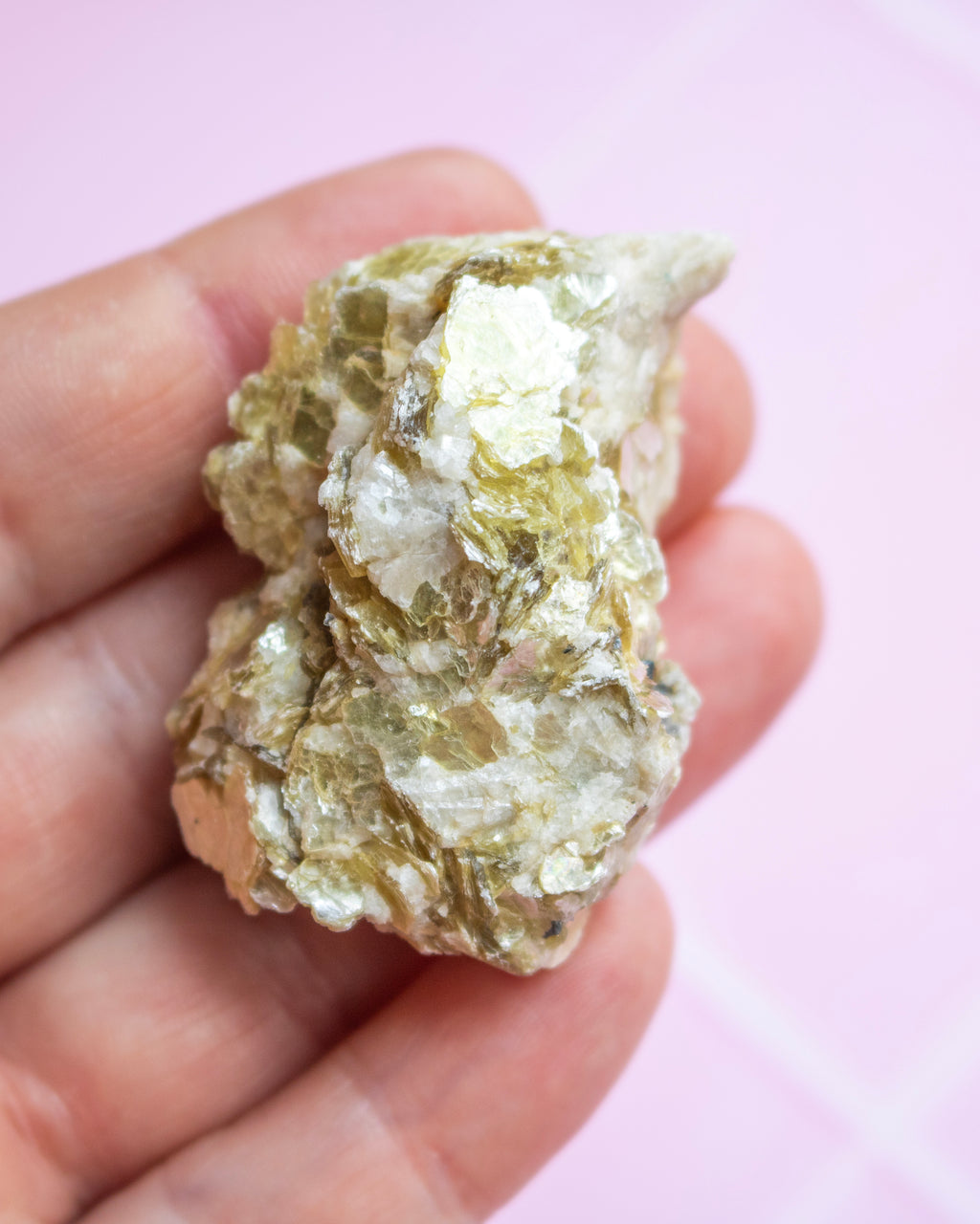 A rough, metallic gold-colored mineral specimen held in a person's hand against a soft pink background.