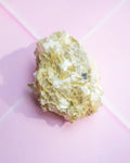 A raw, rough crystal specimen with a mix of pale yellow, cream, and white tones, resting on a pink background with a soft shadow.