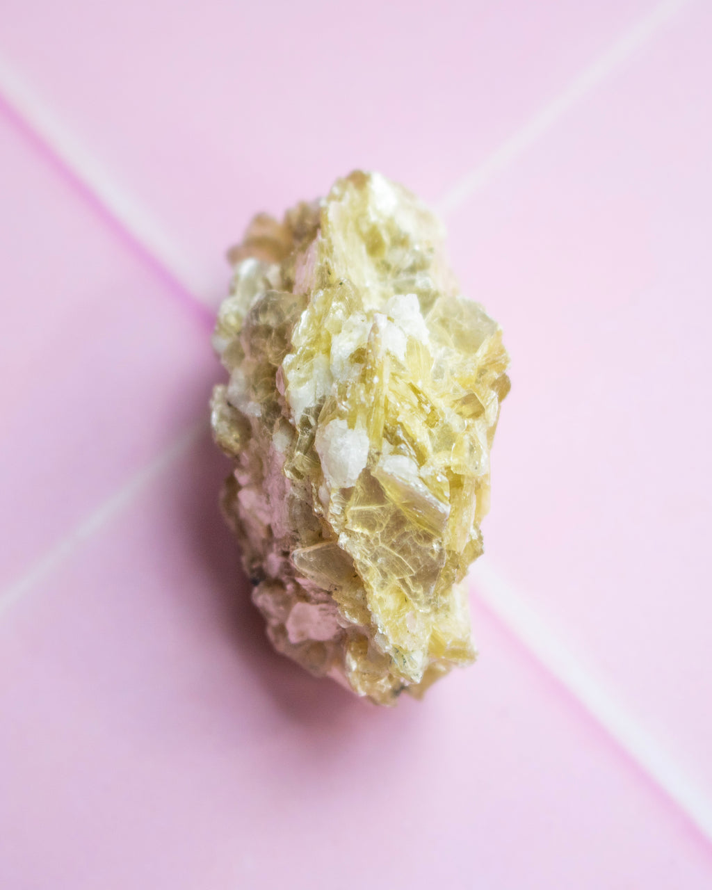 A rough, textured crystal specimen with pale yellow and white crystalline formations, photographed on a soft pink background.
