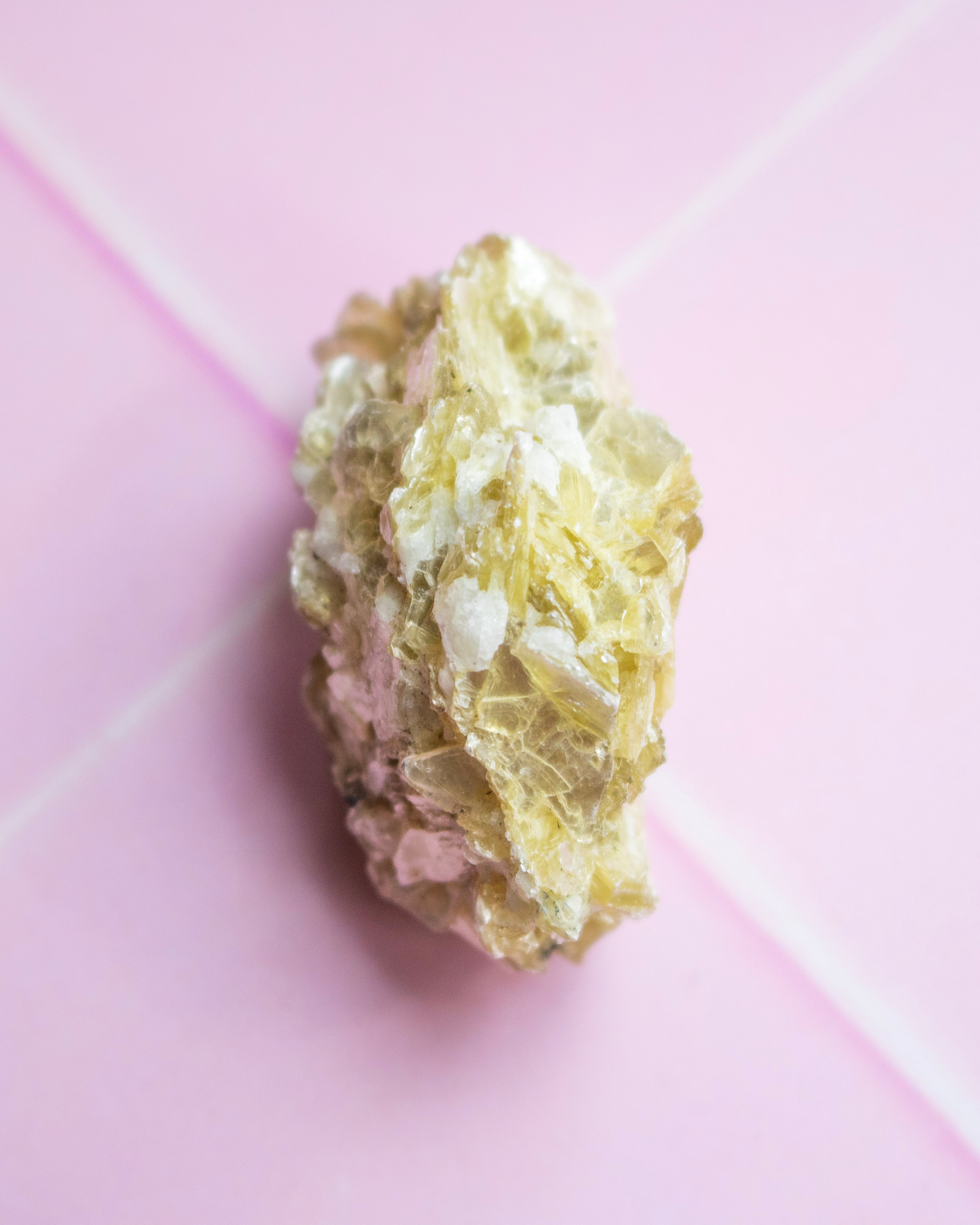 A rough, textured crystal specimen with pale yellow and white crystalline formations, photographed on a soft pink background.