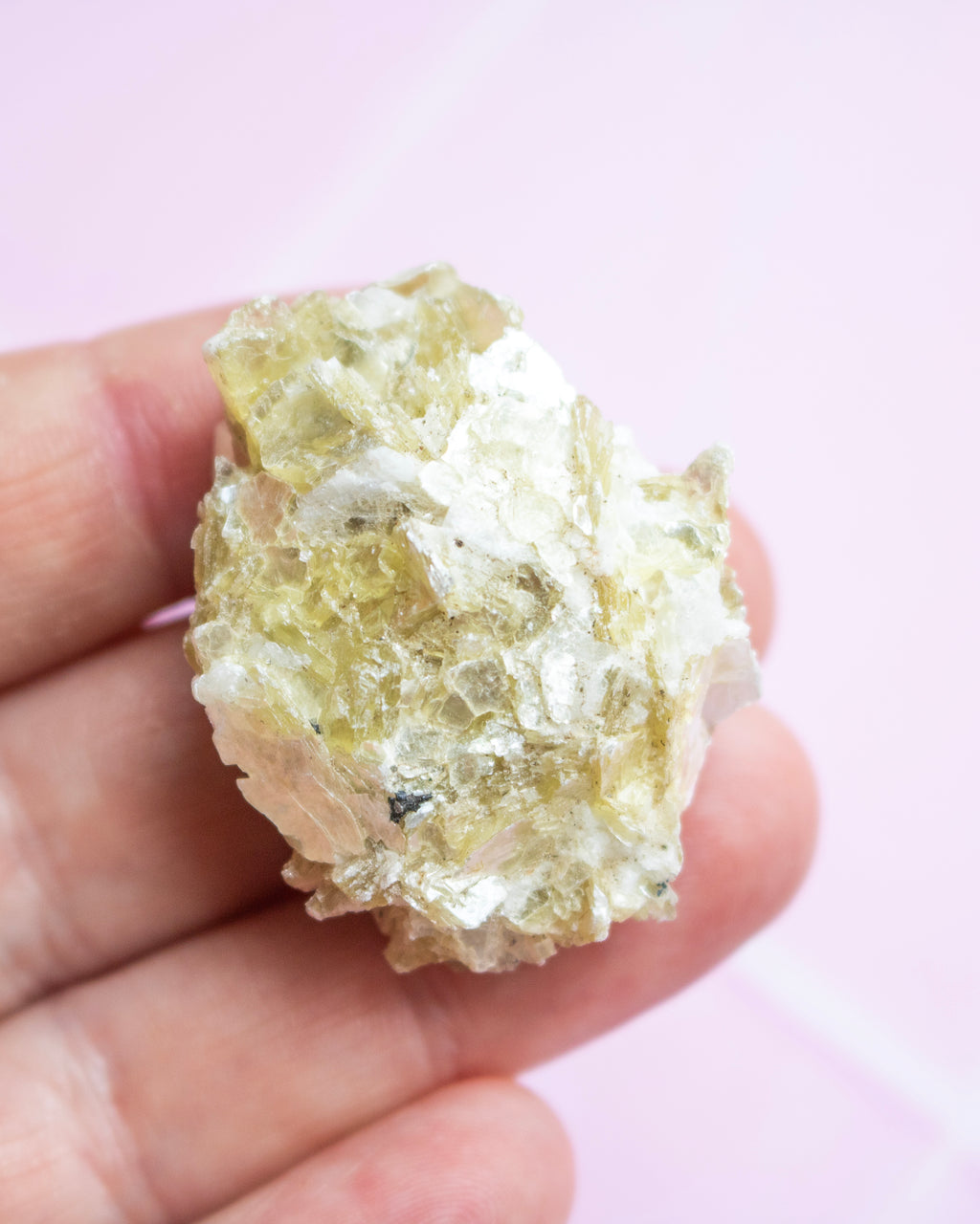 A hand holding a rough, crystalline mineral specimen with pale yellow and translucent white tones against a soft pink background.