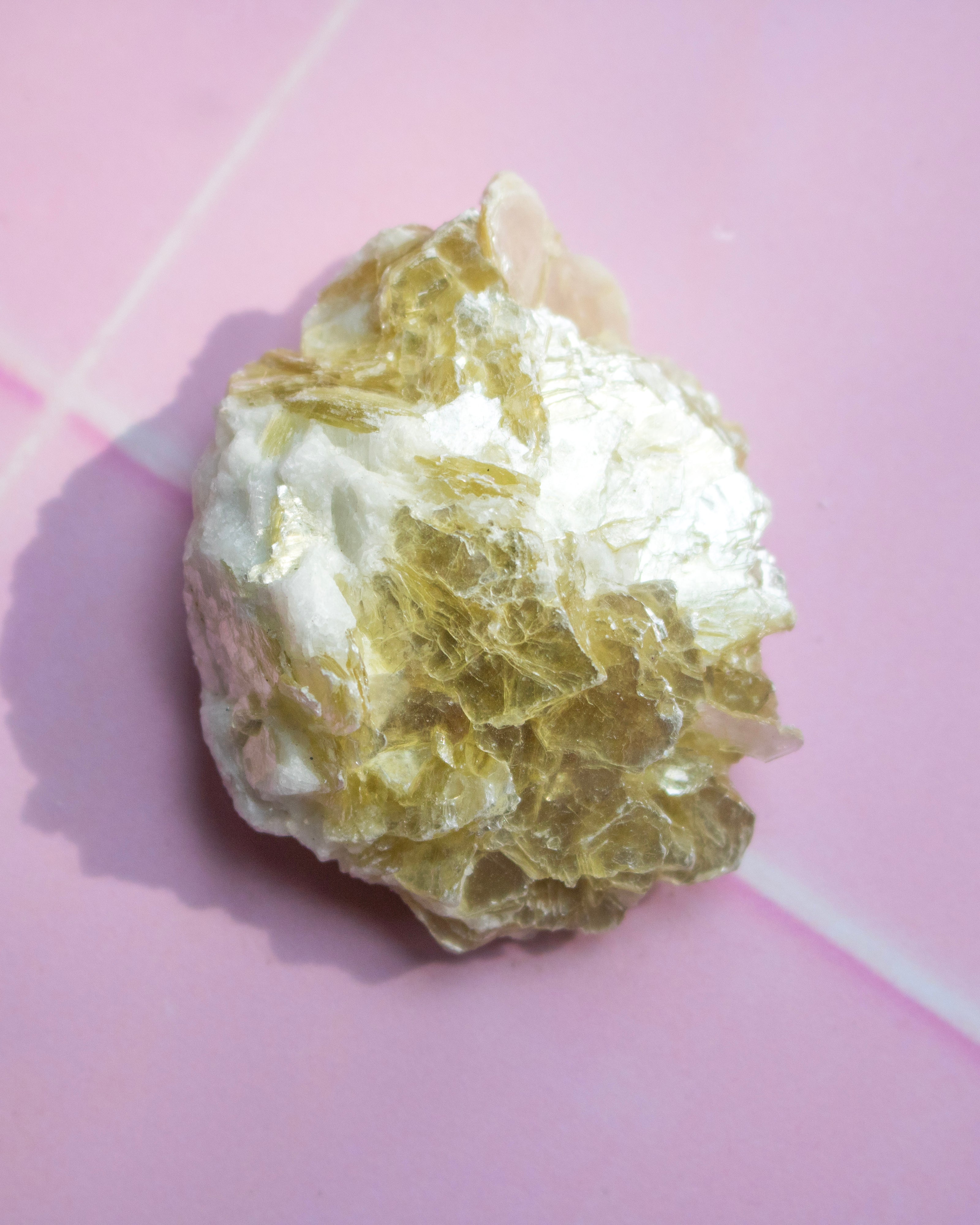 A raw, rough crystal specimen with a mix of white and translucent golden-yellow tones, resting on a pink background.