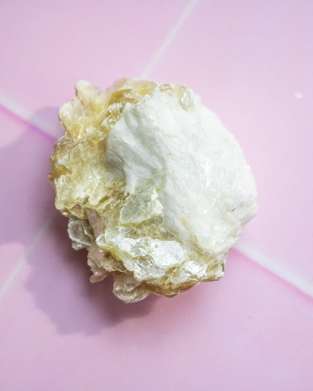 A rough, textured mineral specimen with a mix of white and pale gold crystalline sections, photographed on a pink background.