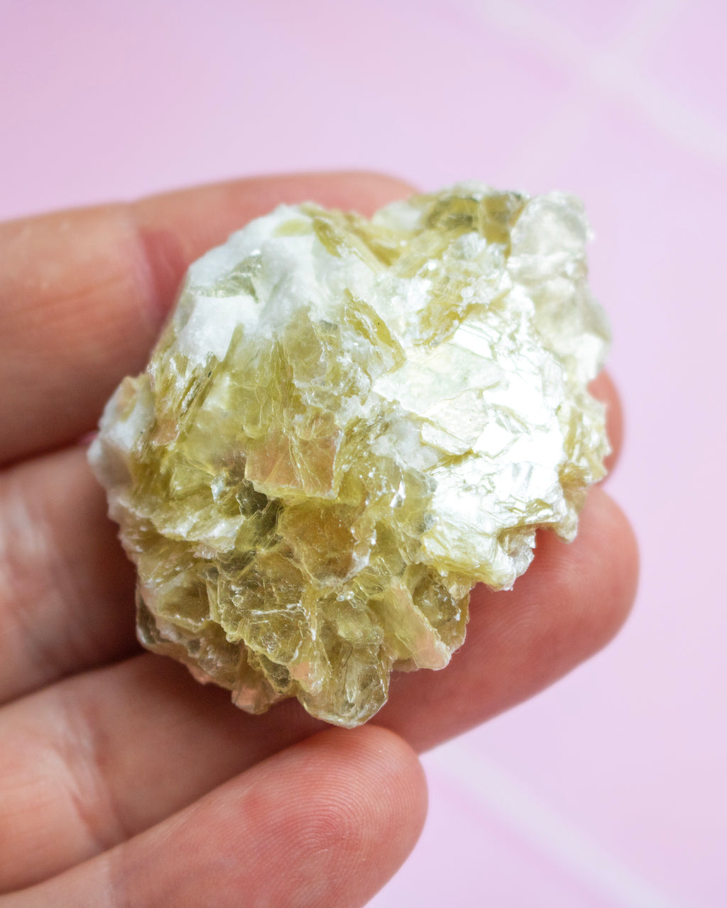 A hand holding a raw, crystalline mineral specimen with pale yellow-green and white tones against a soft pink background.
