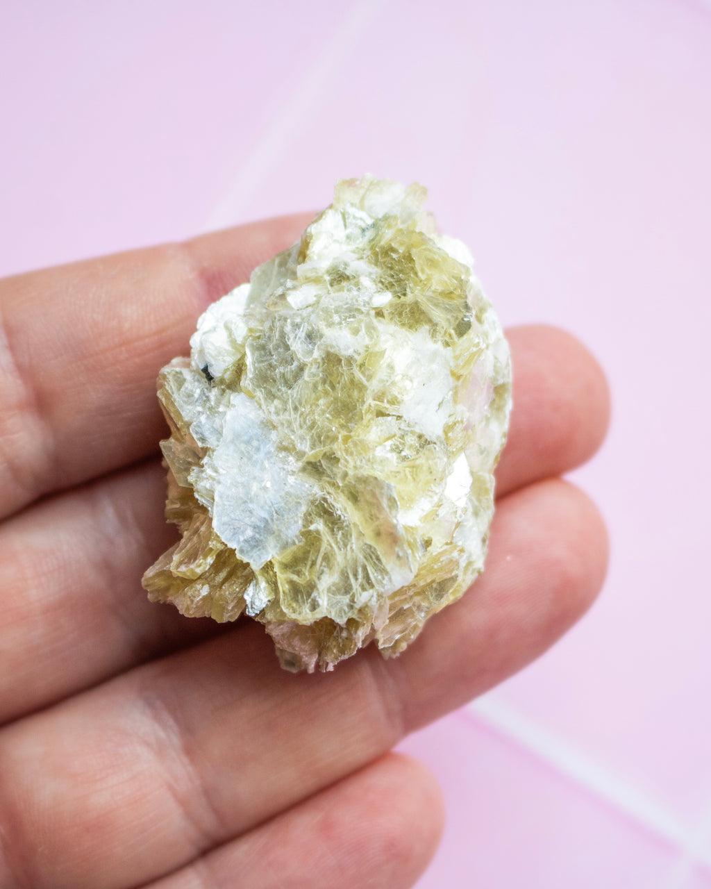 A raw, rough mineral specimen with pale yellow-green and white crystalline texture, held in a person's hand against a soft pink background.