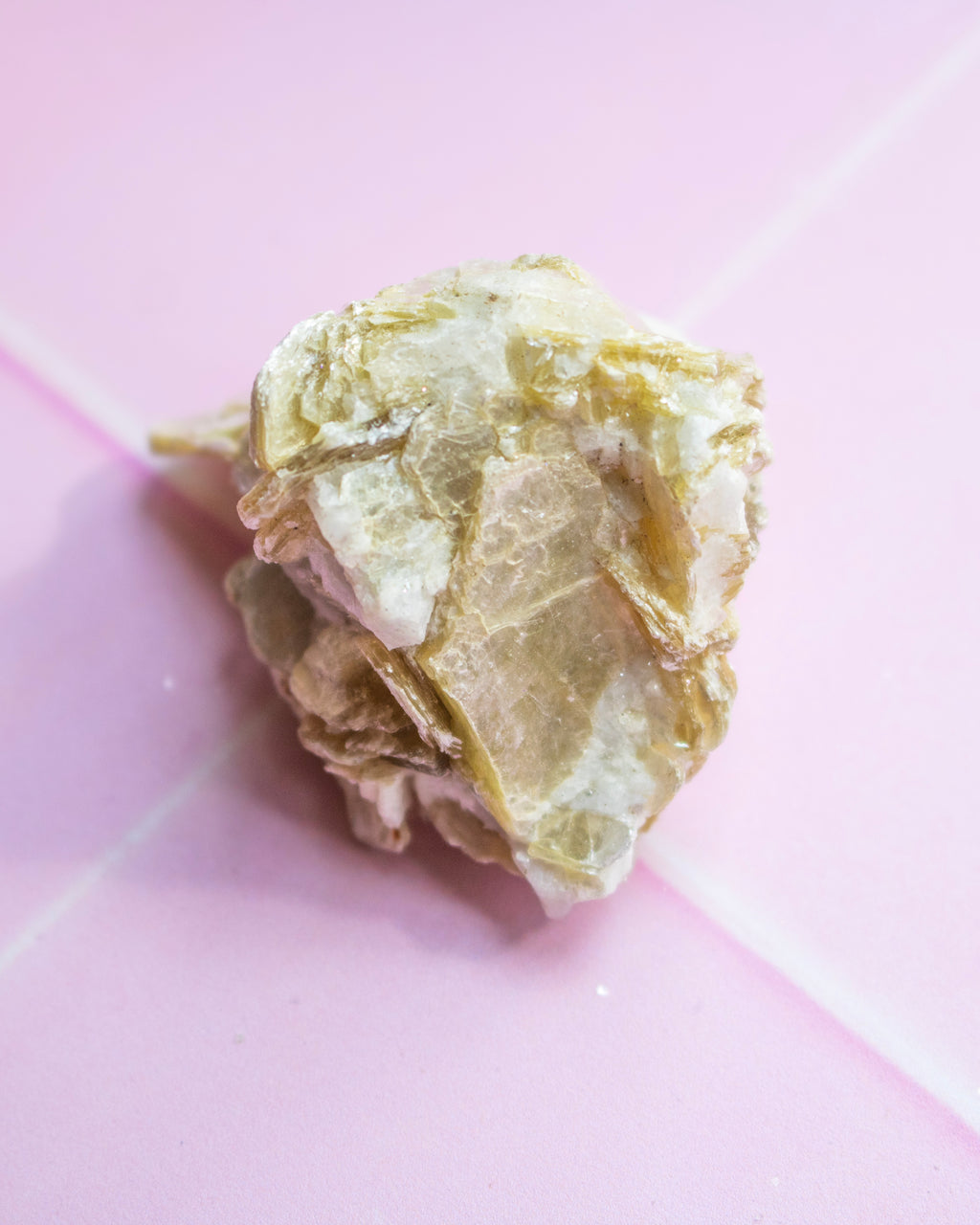 A rough, crystalline mineral specimen with pale gold and off-white tones, photographed on a pink background.