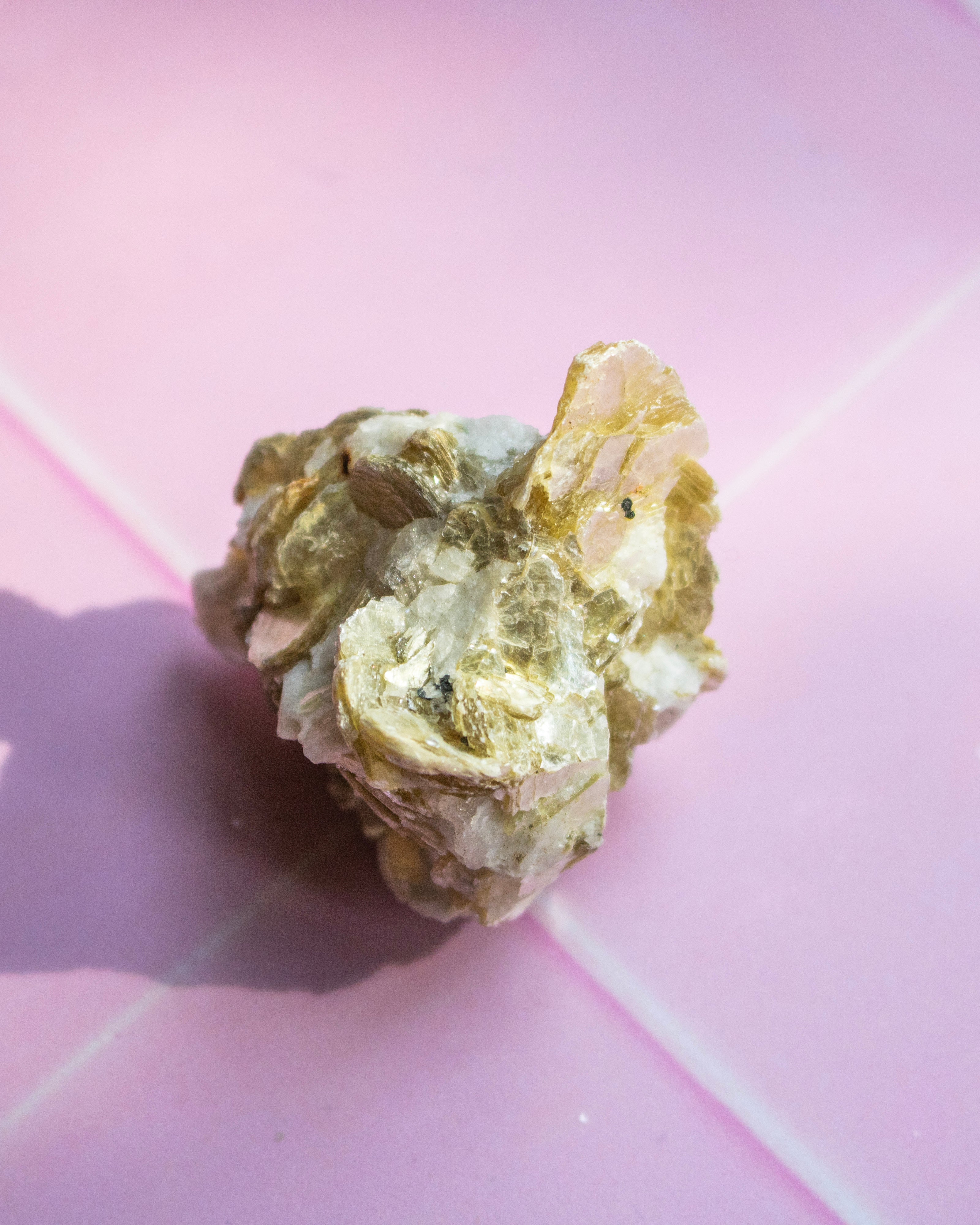 A rough, textured mineral or crystal specimen with metallic gold and pale cream tones, photographed on a pink background.
