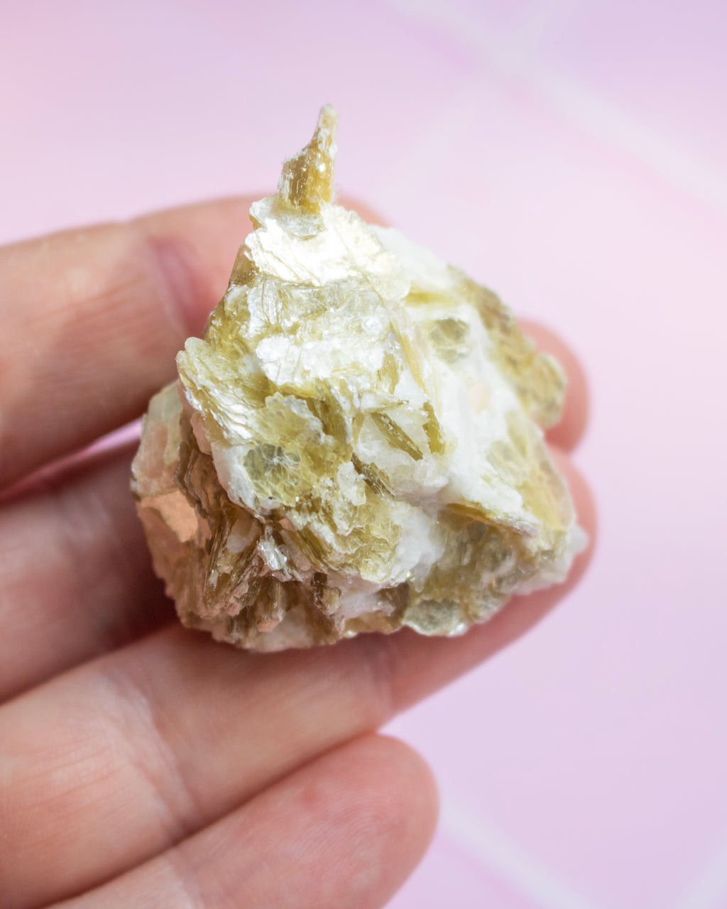 A hand holding a small, rough, pale yellow and off-white mineral or crystal specimen against a soft pink background.