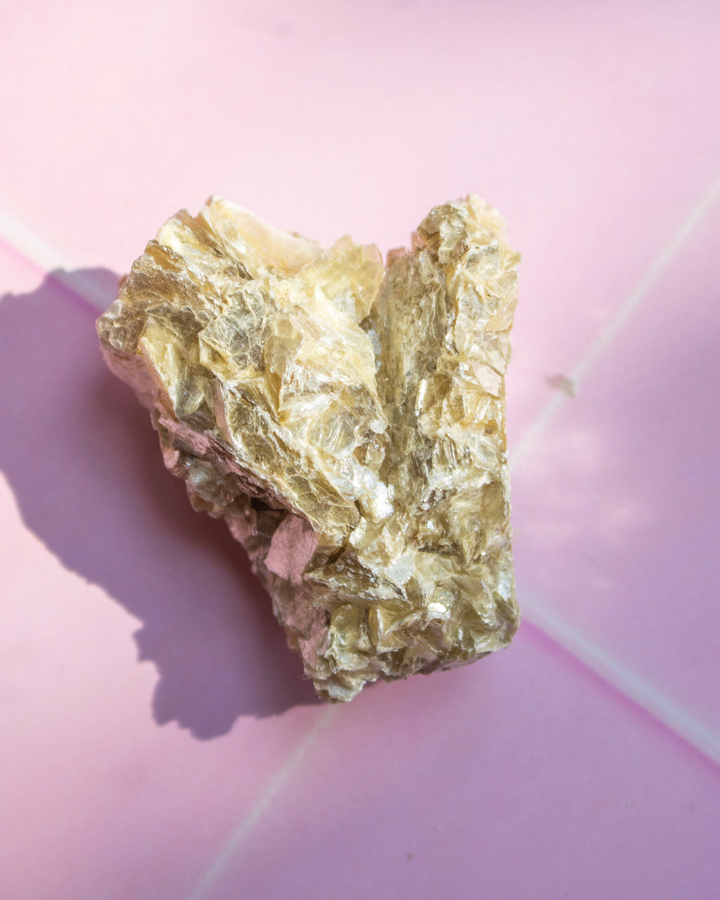 A raw metallic gold crystal specimen with rough, textured flaky surfaces, resting on a pink background with a soft shadow.