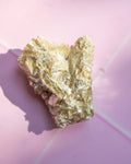 A raw metallic gold crystal specimen with rough, textured flaky surfaces, resting on a pink background with a soft shadow.
