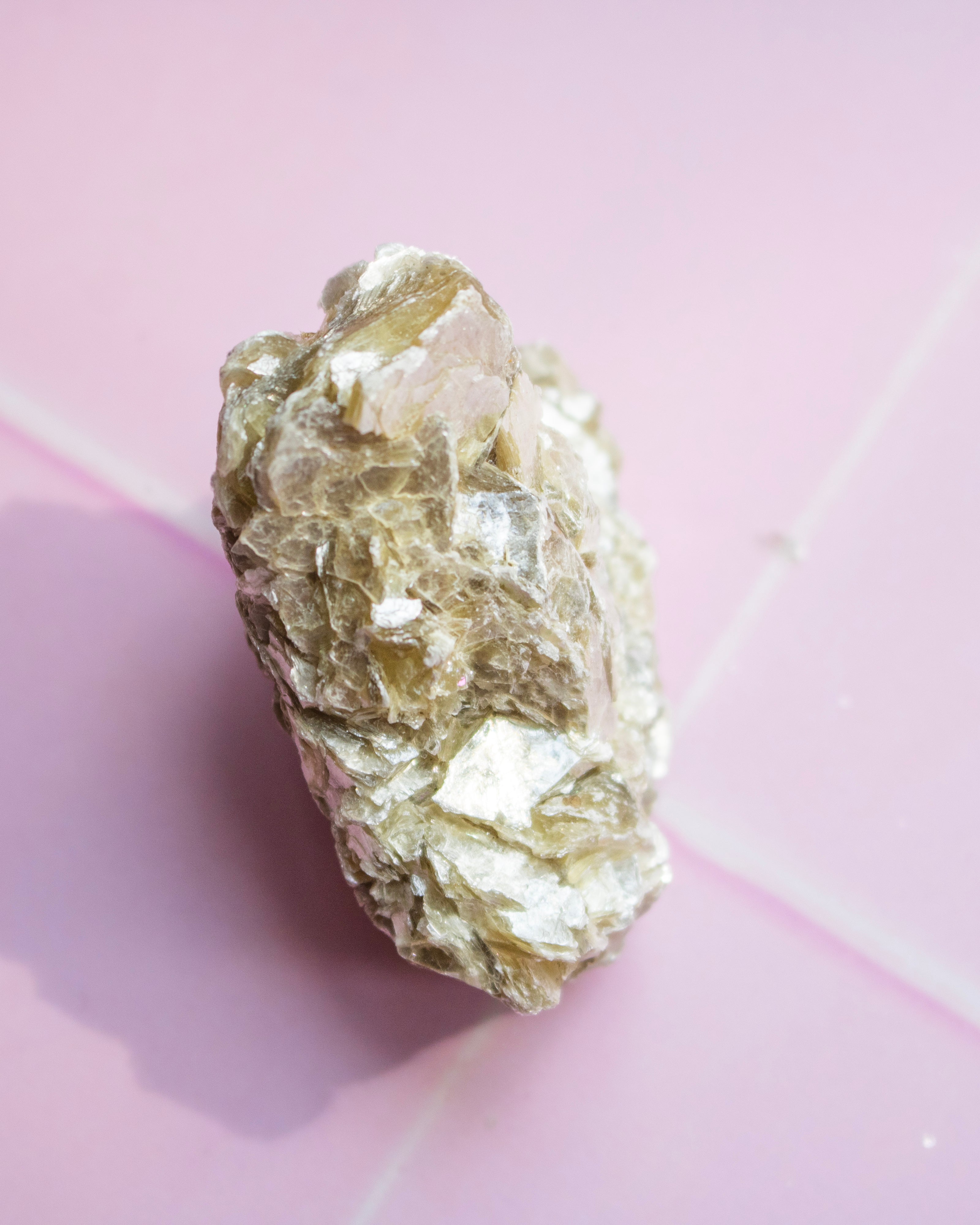 A rough, metallic gold-colored mineral specimen with a crystalline texture, photographed on a soft pink background.