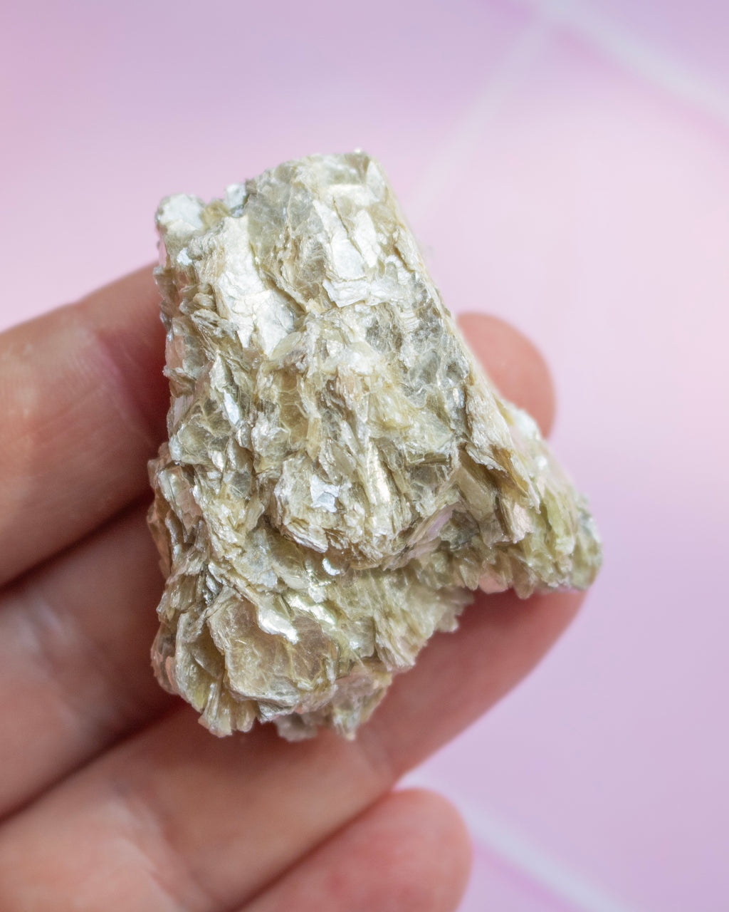 A hand holding a rough, metallic silver-colored mineral or crystal specimen against a soft pink background.