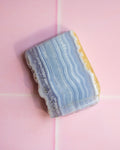 A polished rectangular stone slab with wavy light blue, white, and gray bands and a yellow-gold edge, photographed on a pink background.