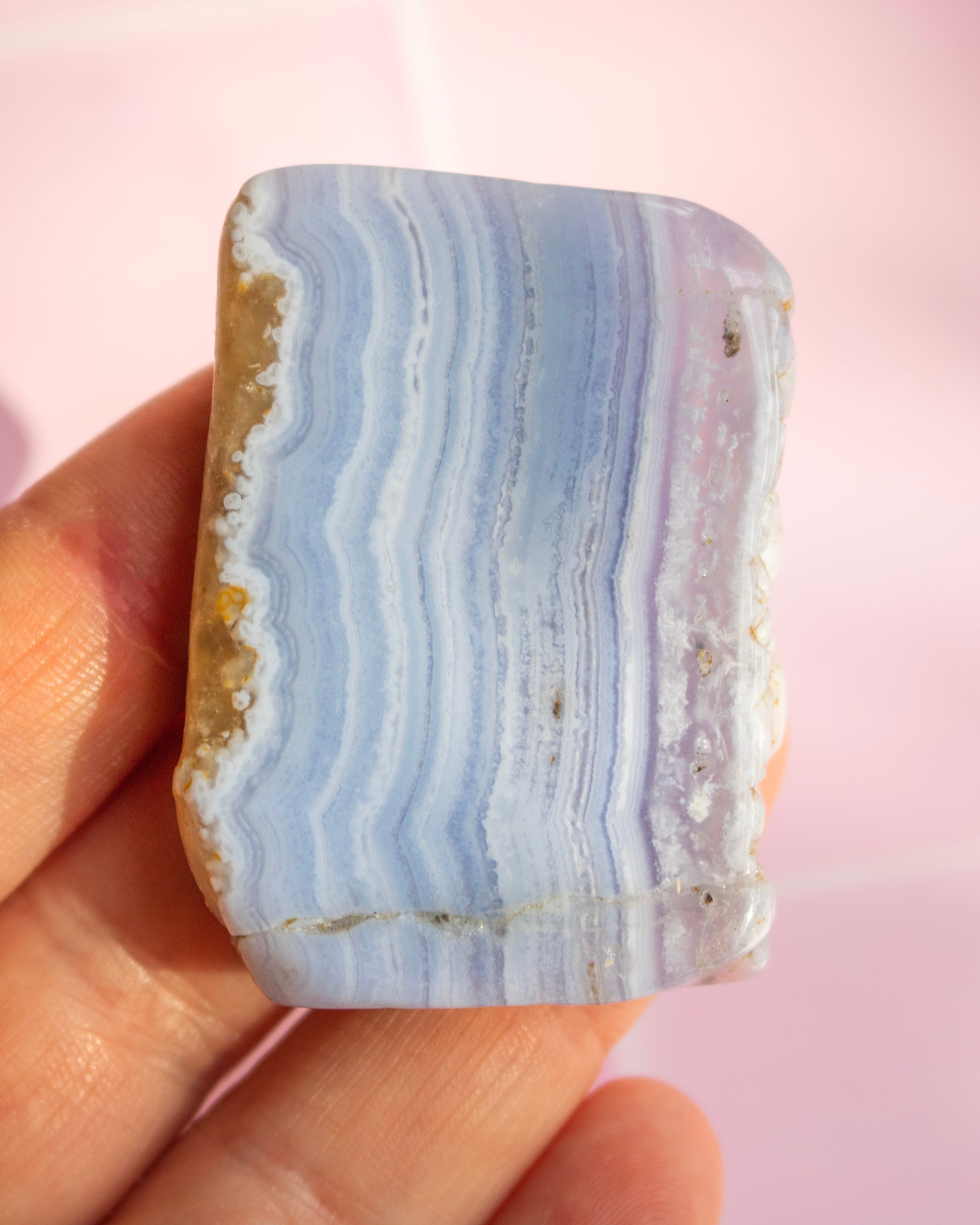 A hand holding a polished rectangular stone with wavy light blue, white, and lavender-gray banded stripes, showing a rough tan matrix on one side, against a soft pink background.
