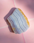 A polished stone slice with layered white, pale blue, and lavender bands and a rough golden-brown textured edge, photographed on a pink surface.
