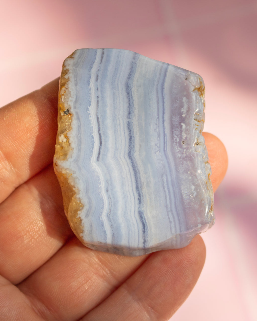 A hand holding a polished natural agate stone with layered light blue, lavender, and white bands, showing rough tan matrix on one side, against a soft pink background.