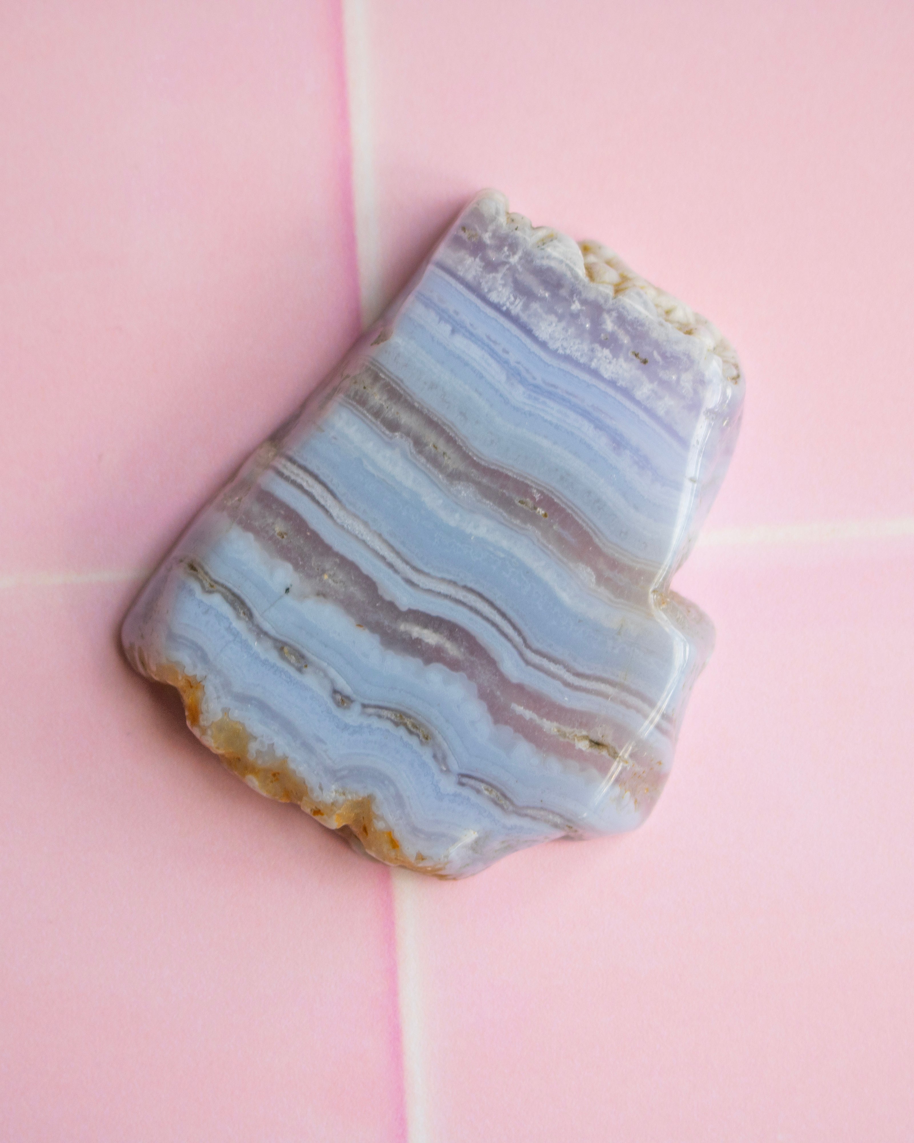 A polished agate stone slab with wavy bands of pale blue, lavender, gray, and tan, photographed on a pink background.