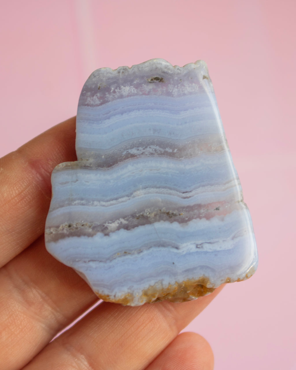 A hand holding a polished agate stone slice with horizontal bands of pale blue, lavender, and white, set against a solid pink background.