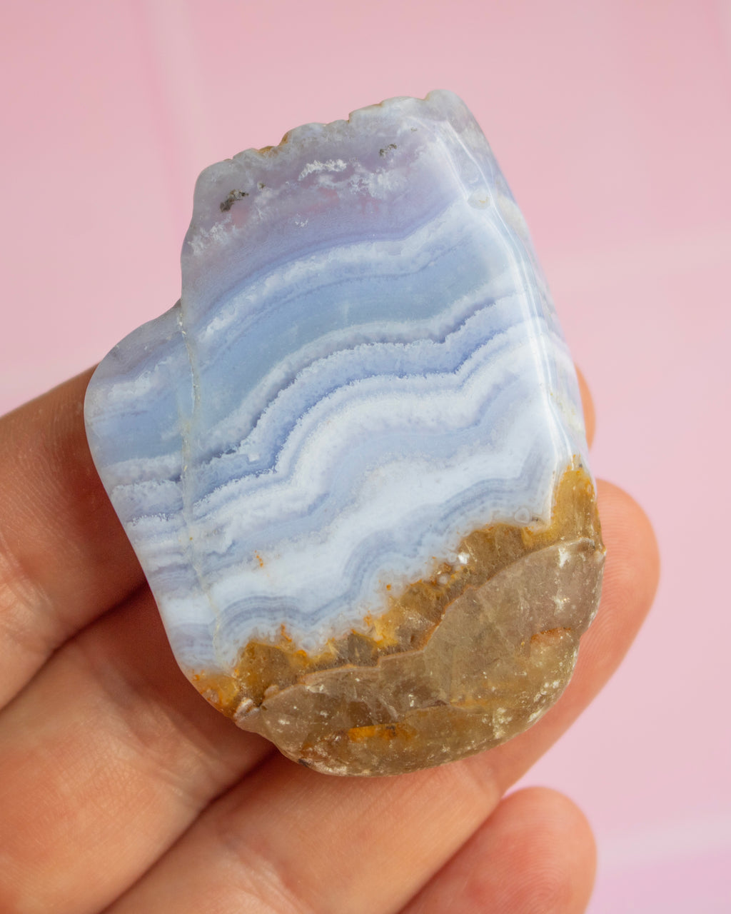 A hand holding a polished natural stone with layered blue, white, and lavender bands and a brown crystalline base, set against a soft pink background.