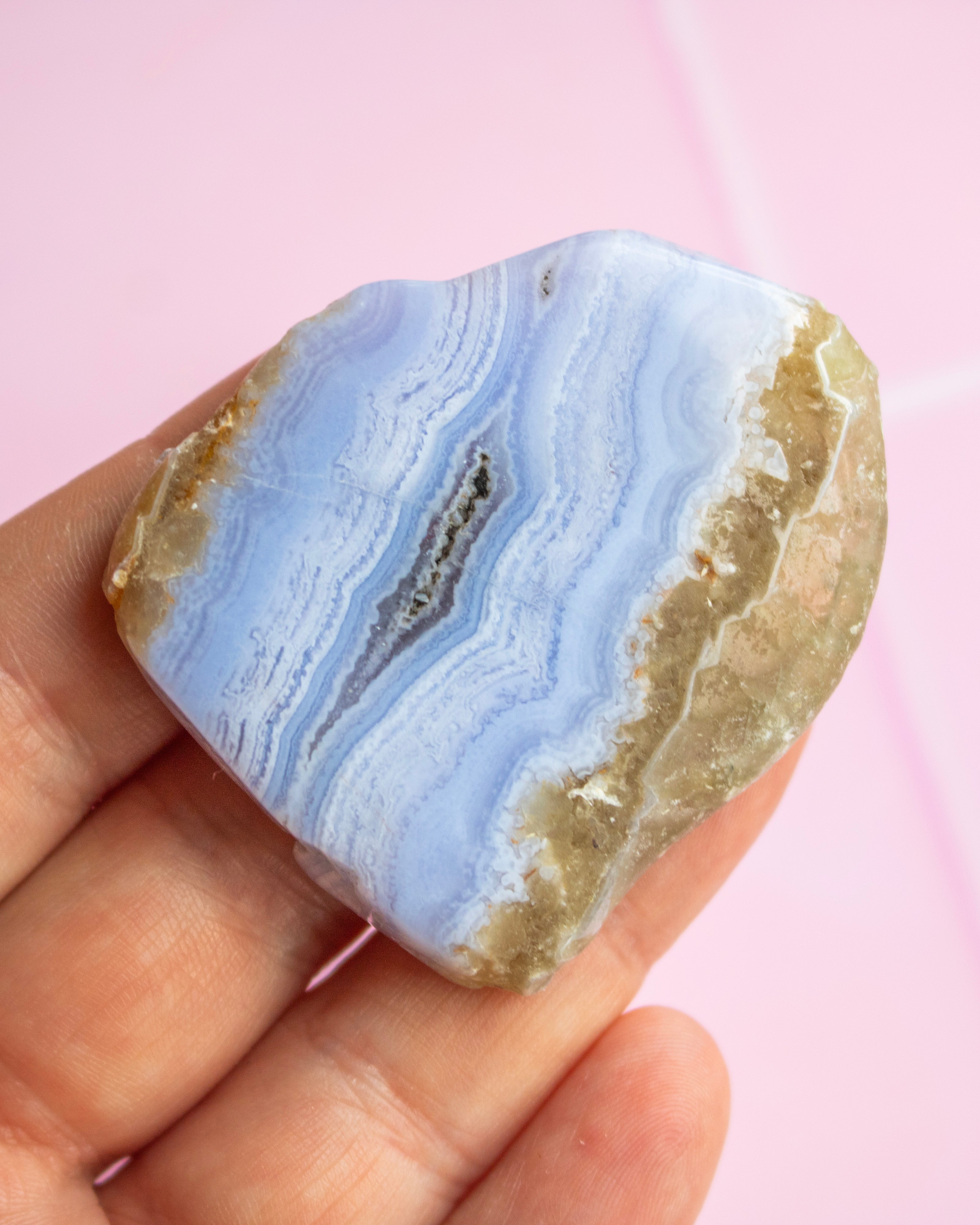 A hand holding a polished natural agate stone with light blue banded bands and rough tan edges, set against a soft pink background.